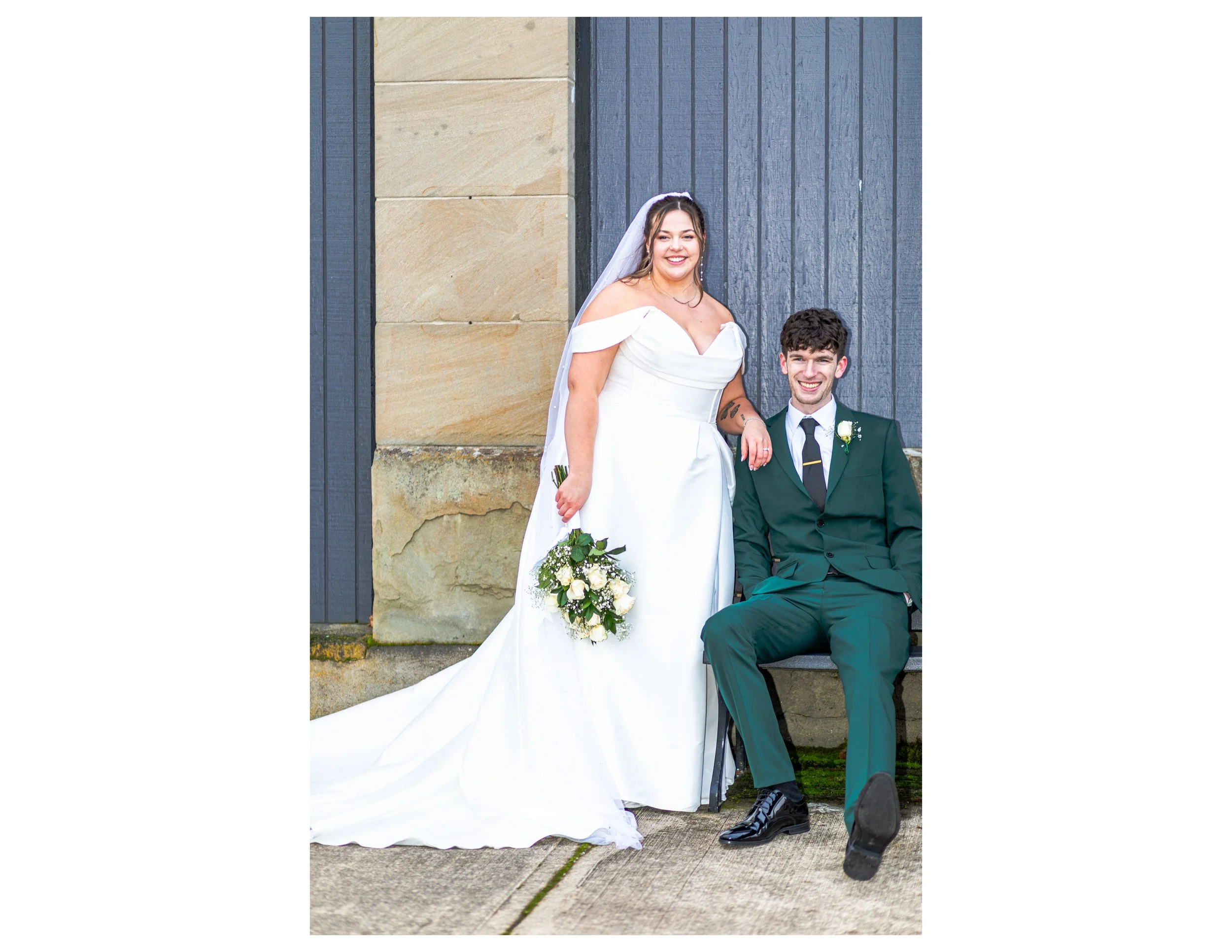 A bride and groom posing outdoors against a building with a blue wooden and beige stone wall. The bride is in a white off-the-shoulder wedding dress with a long train, and the groom is in a green suit with a white shirt and black tie. The bride is ho