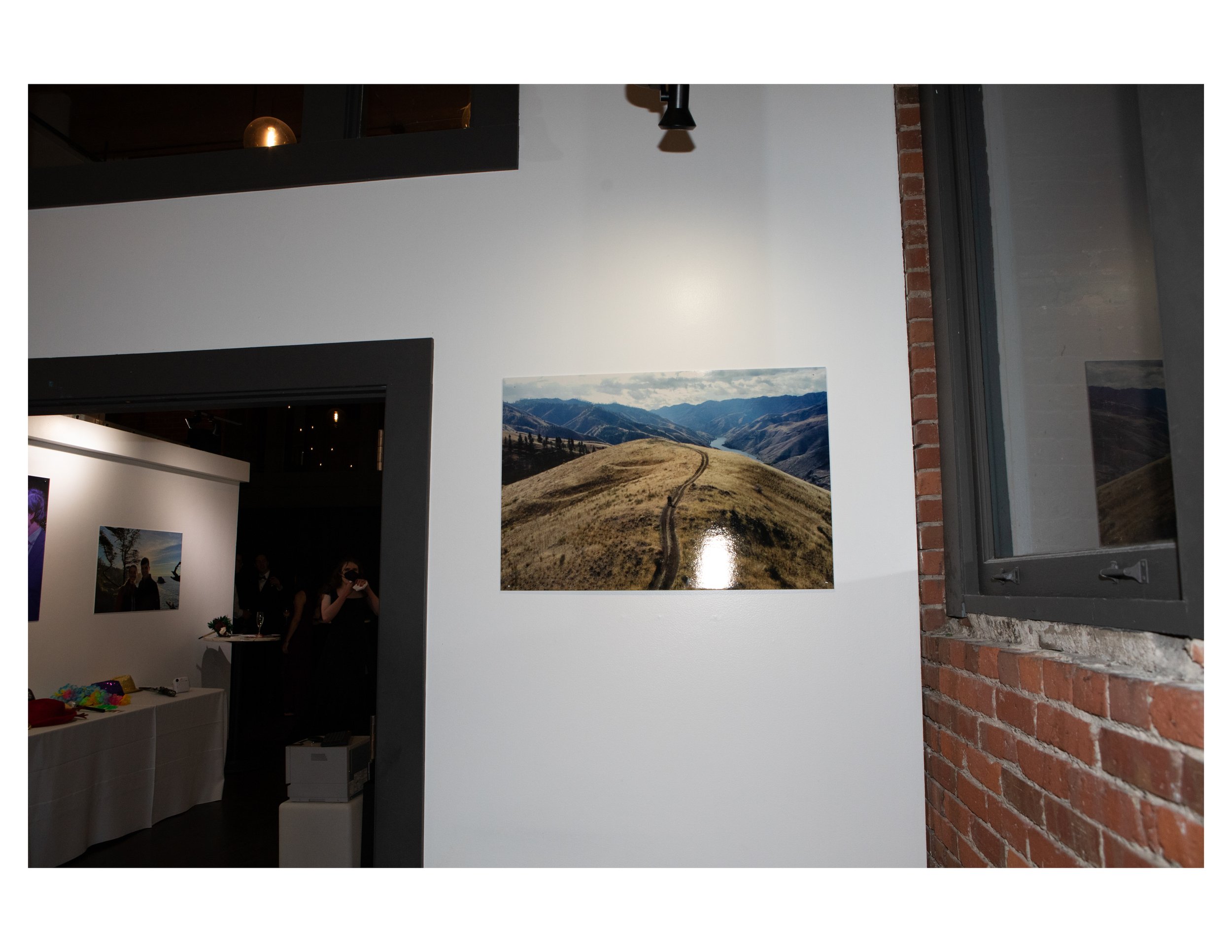 Interior of an art gallery with paintings on white walls, a brick wall on the right, and a window on the right side. There is a group of people in the background, and a table with miscellaneous items in the foreground.