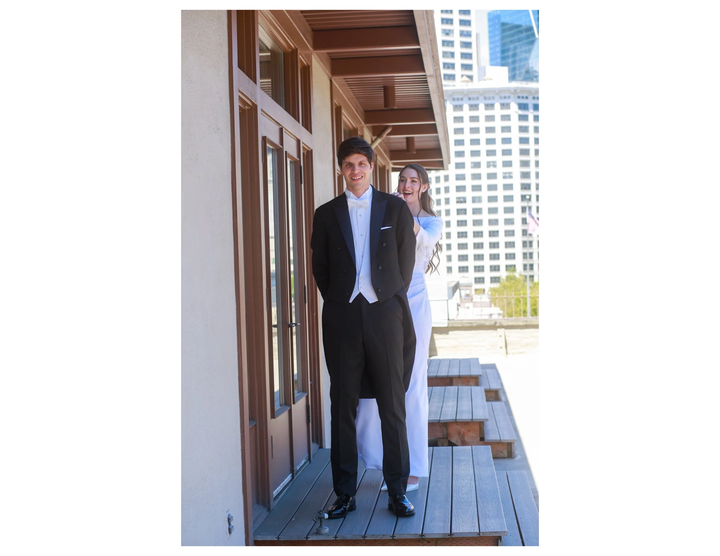 A man in formal attire, a tuxedo with a bow tie, and a woman in a white dress are standing on a balcony in an urban area with tall buildings in the background. The man is smiling and facing forward, while the woman is slightly behind him, smiling and
