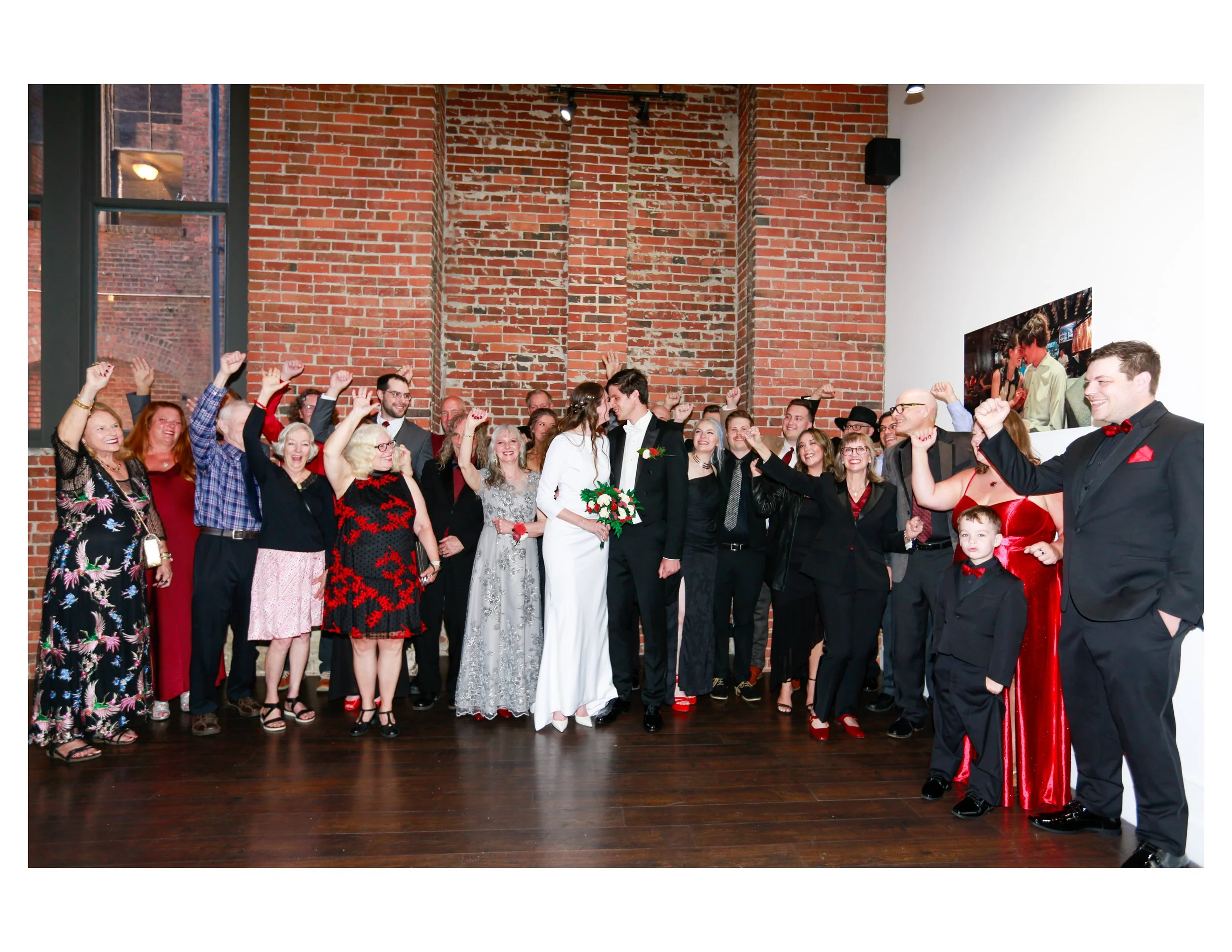 A group of people dressed in formal attire celebrating at a wedding reception, with some raising their hands and smiling, in front of a brick wall.