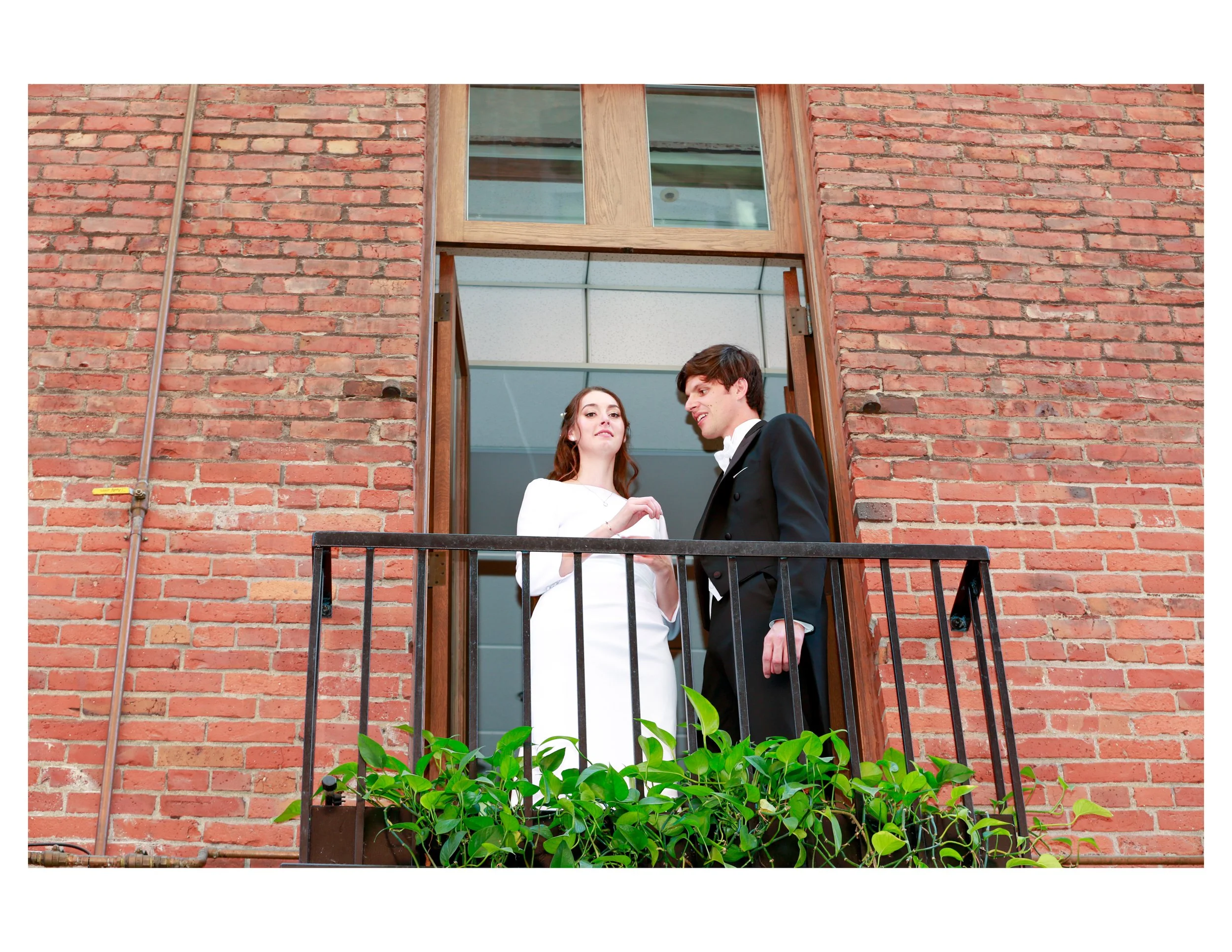 A bride and groom standing on a balcony in front of a brick building, with the bride wearing a white dress and the groom in a black tuxedo, looking at each other.