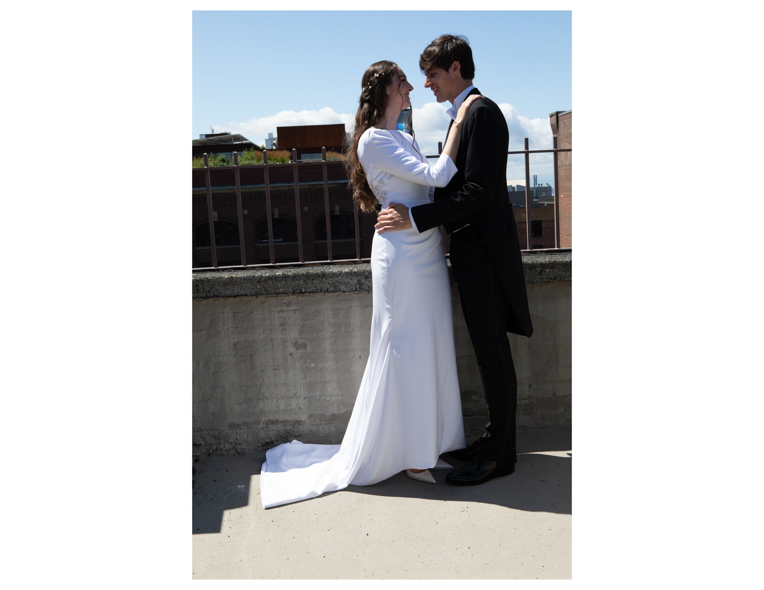 A bride and groom are standing close together on a rooftop, smiling and looking into each other's eyes. The bride is wearing a white wedding dress, and the groom is dressed in a black suit. They are embracing each other with a cityscape and partly cl