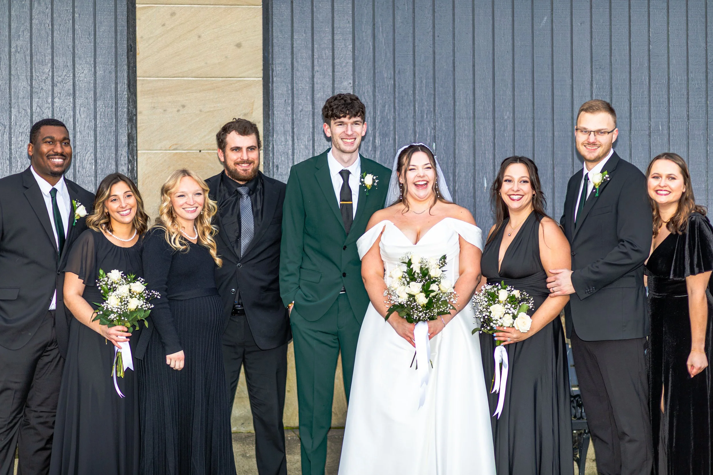 Group of people at a wedding celebration, with brides and grooms in formal attire, smiling and holding bouquets, standing outside against a wooden backdrop.