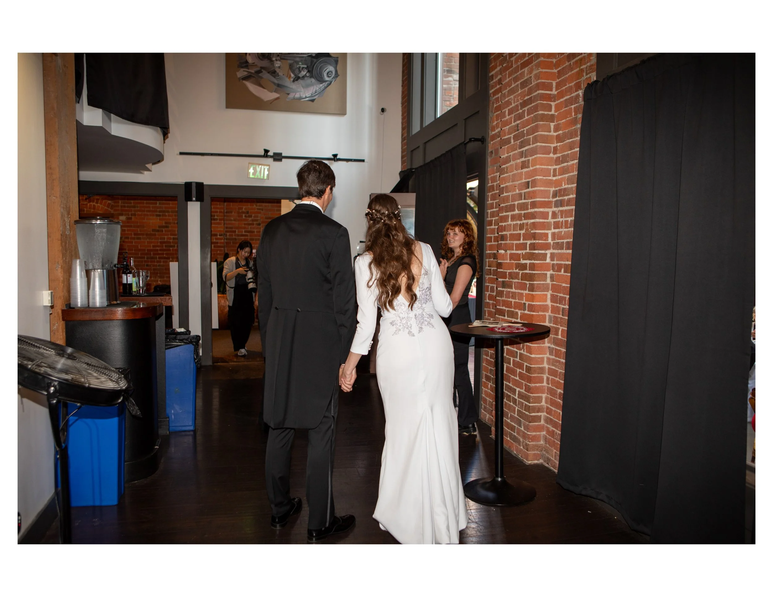 Wedding couple, man in tuxedo and woman in white gown, facing each other in an indoor venue with brick walls.