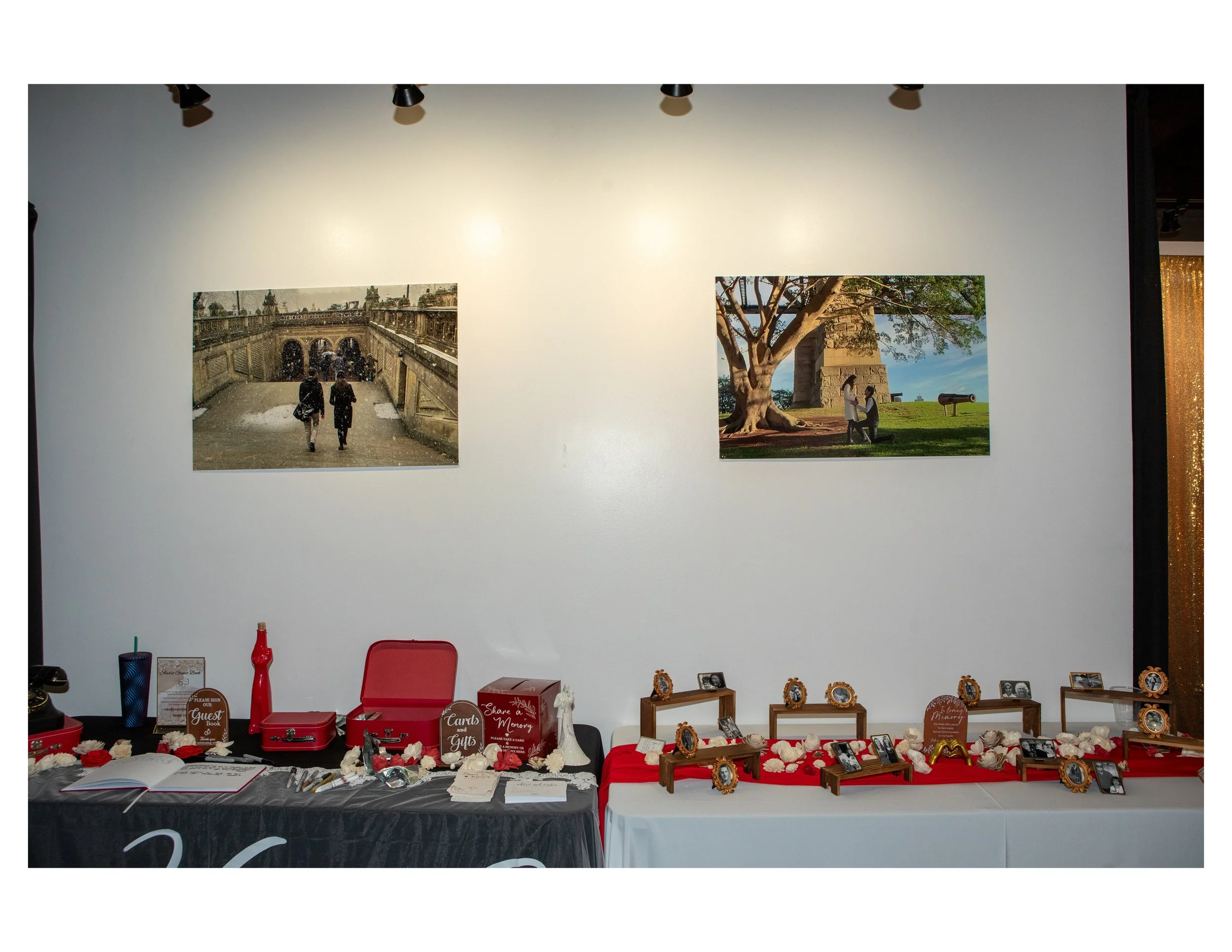 Display table with framed photographs, small decorative items, and signs, set against a wall with two landscape photos hung above, one showing a historical site with two people walking and the other showing a large tree with a couple underneath, in a