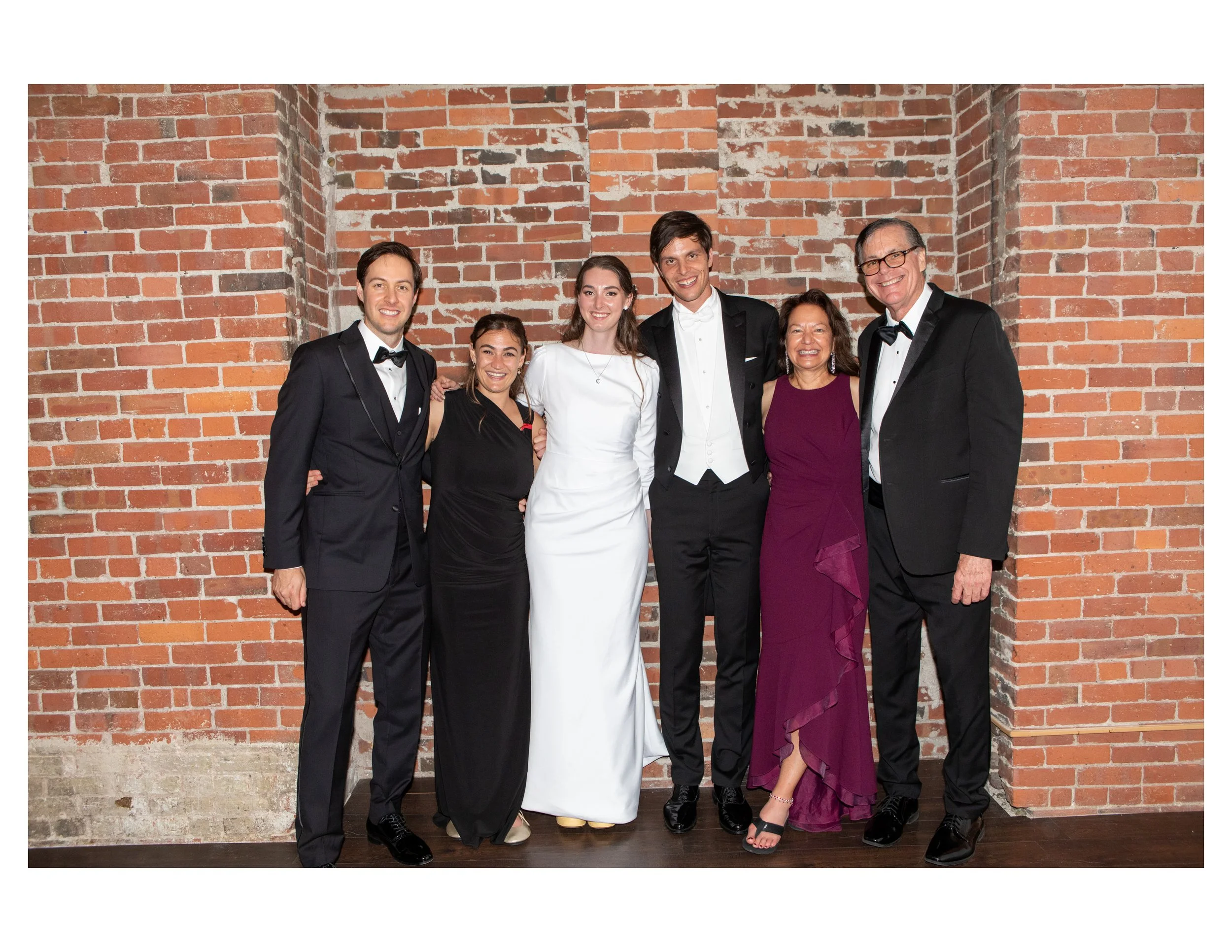 Group of six people dressed in formal clothing standing in front of a brick wall, smiling at the camera.