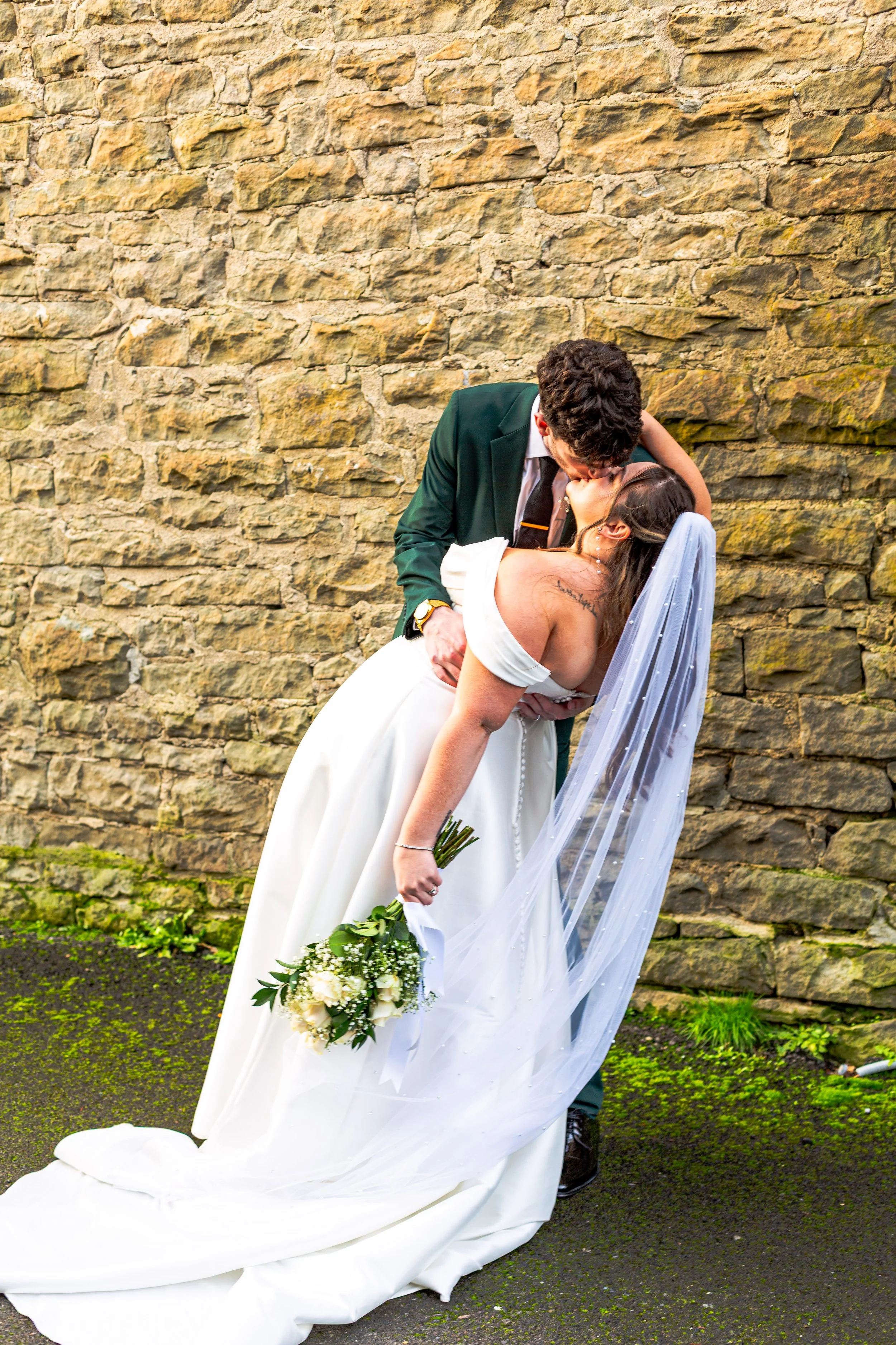 A bride and groom sharing a kiss outside, with the groom holding the bride who is holding a bouquet of white roses, against a stone wall background.