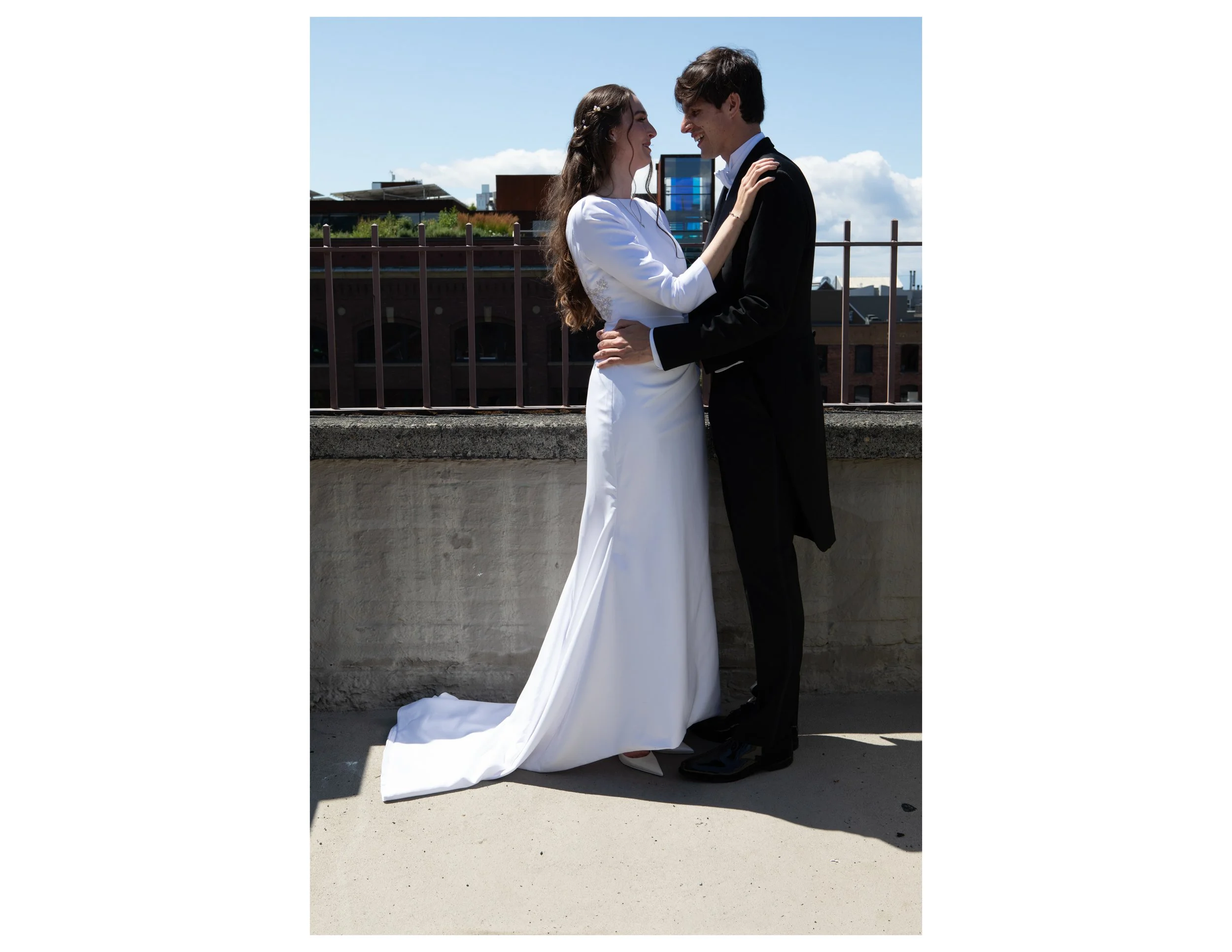 A bride and groom are standing close together on a rooftop, embracing and smiling at each other under a partly cloudy sky.