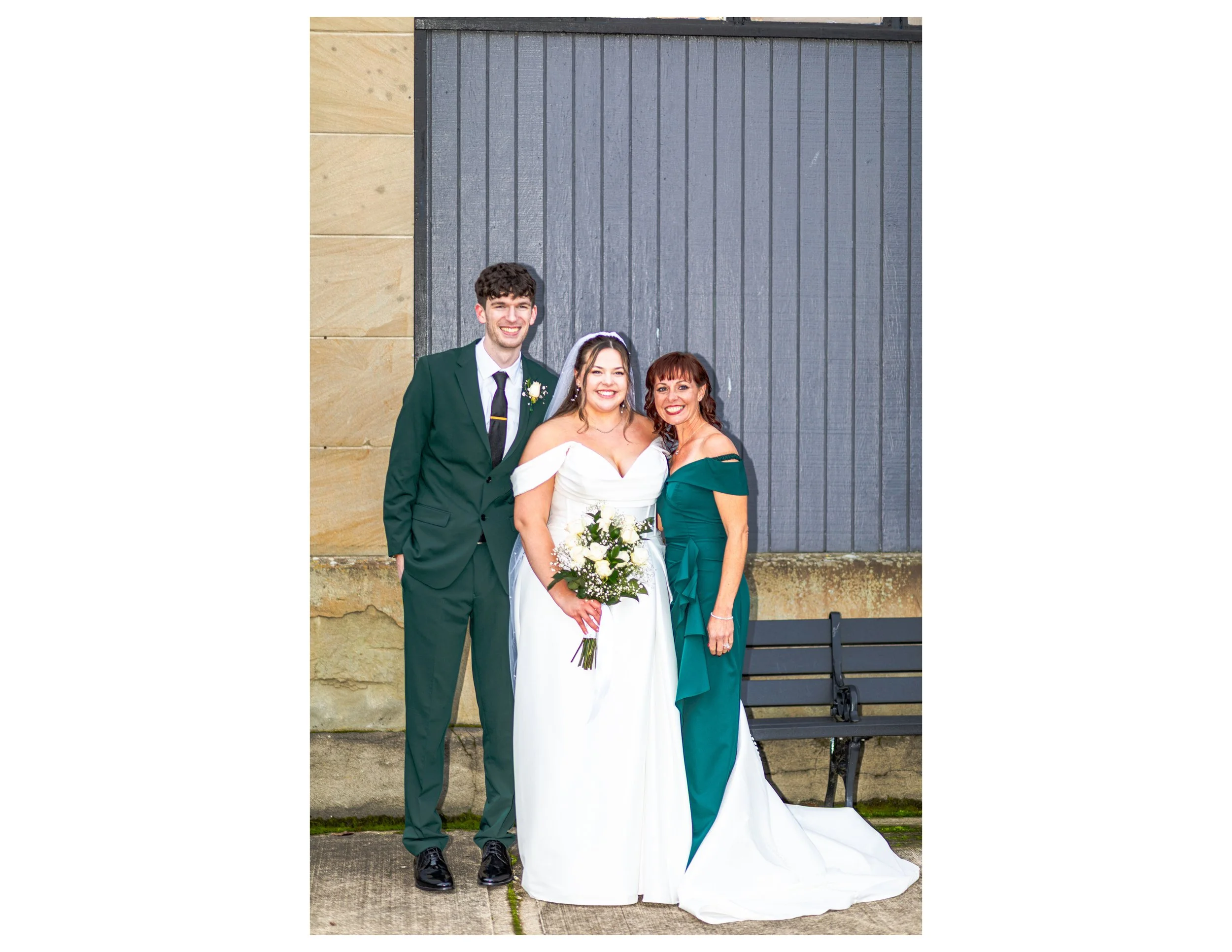 Three people standing in front of a gray door, dressed in formal attire for a wedding. The man is wearing a dark green suit, white shirt, and black tie. The woman in the middle is wearing a white wedding gown and holding a bouquet of flowers. The wom