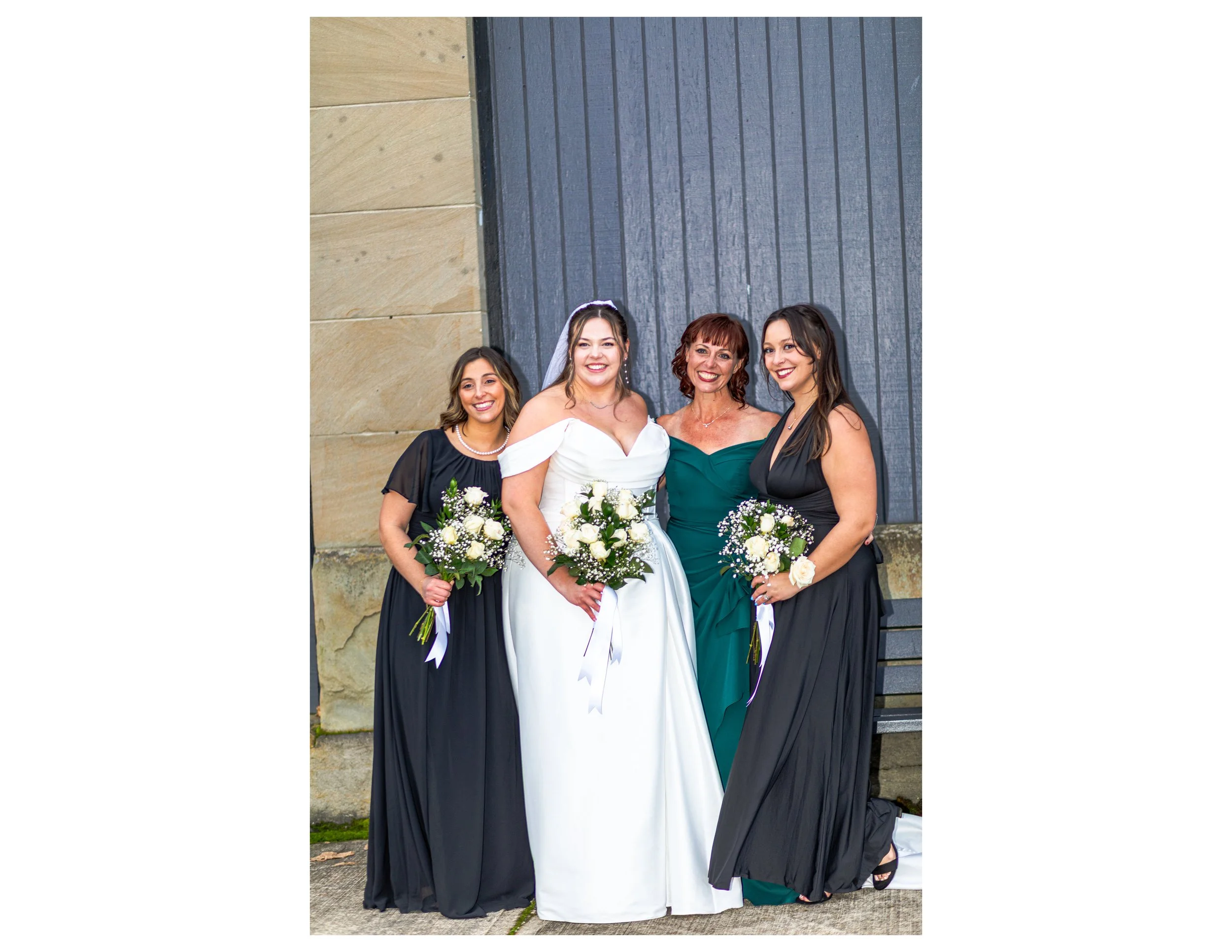 Four women standing outdoors in front of a building with mixed stone and dark wooden siding, dressed in formal attire for a wedding, holding bouquets of white flowers and smiling.