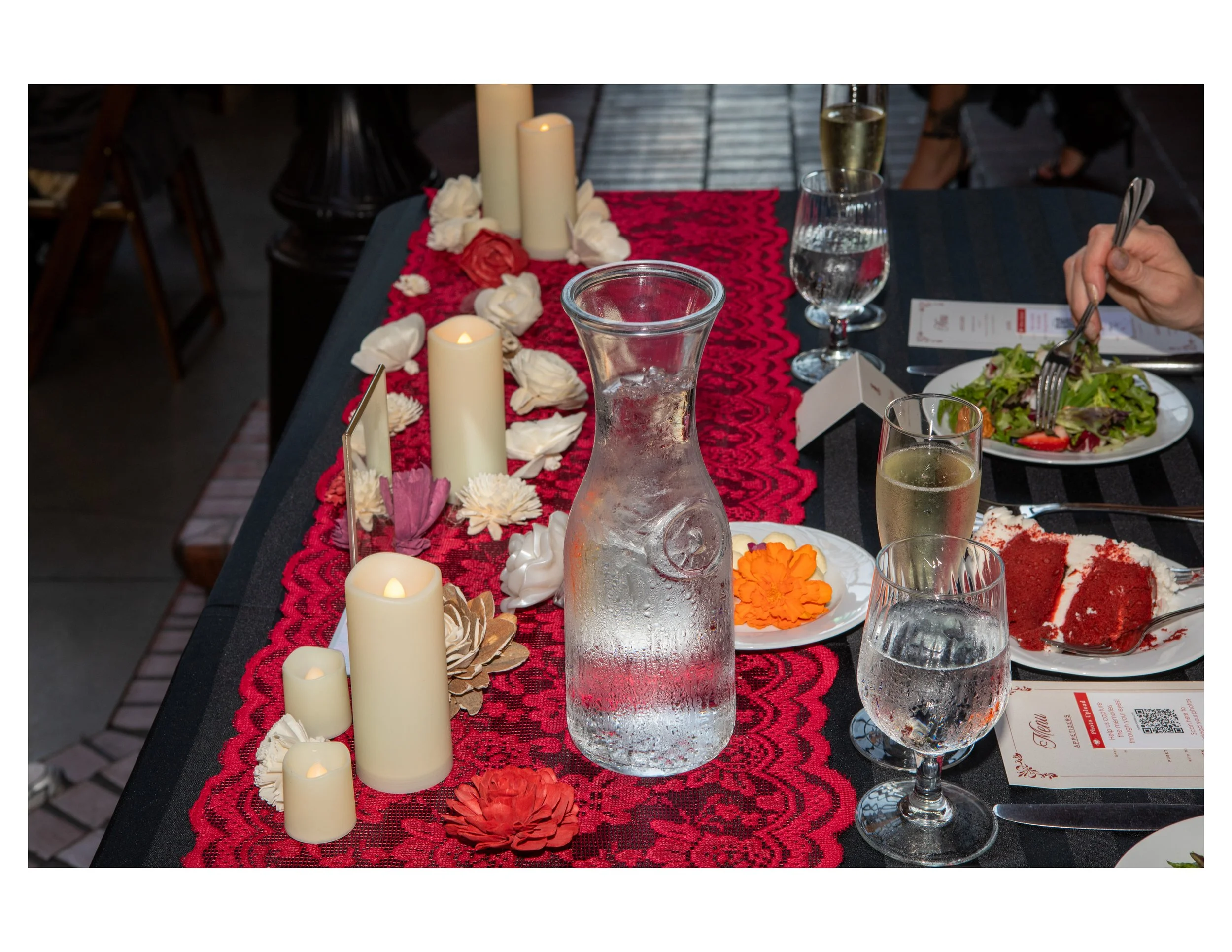 A decorated dining table with white candles, flower petals, candles, glasses of water, plates of food, and a person holding a fork. The table has a red table runner and a black surface.