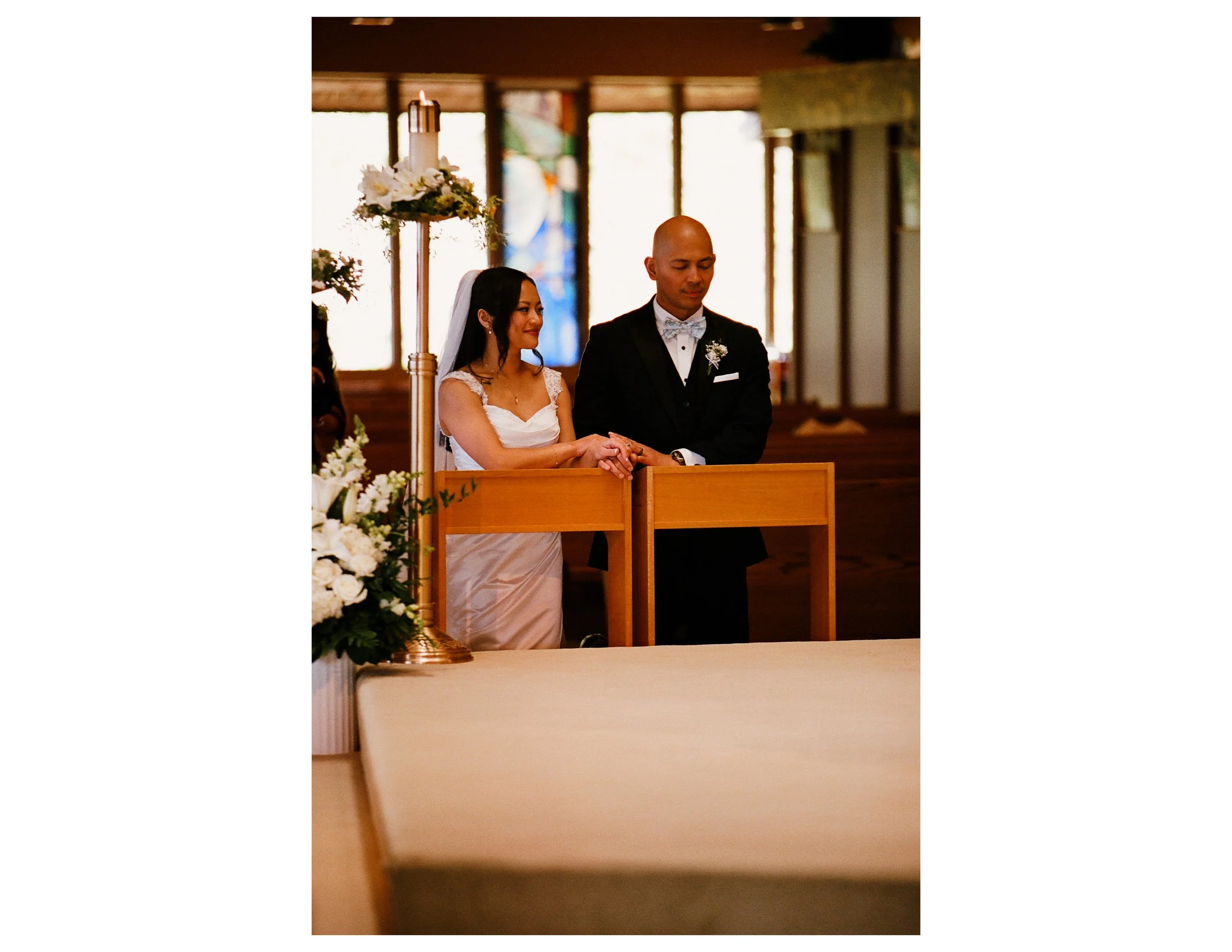 A bride and groom standing side by side during a wedding ceremony inside a church, with a floral arrangement and candle on a stand in the foreground.