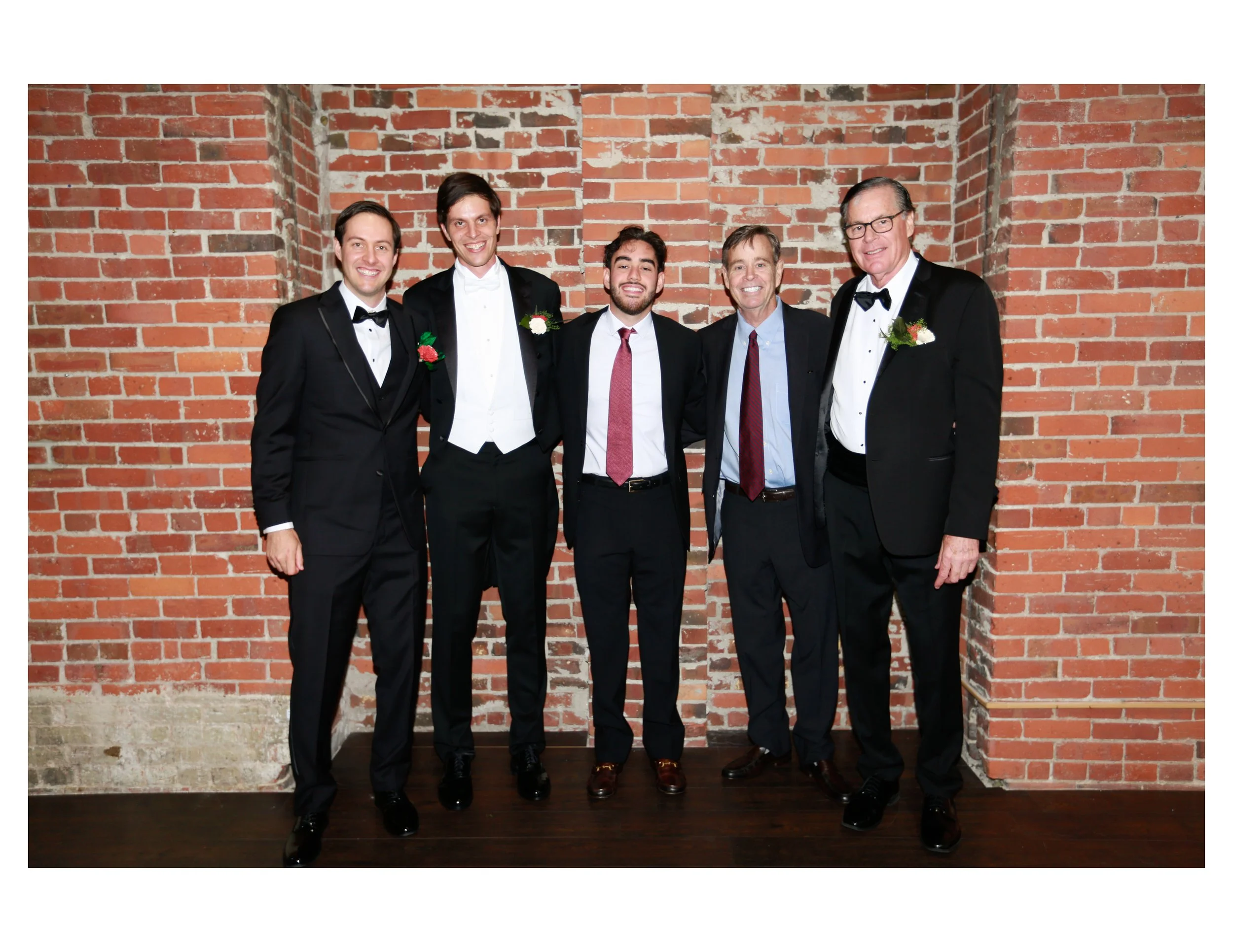 Five men dressed in tuxedos and suits standing together against a brick wall, smiling at the camera.