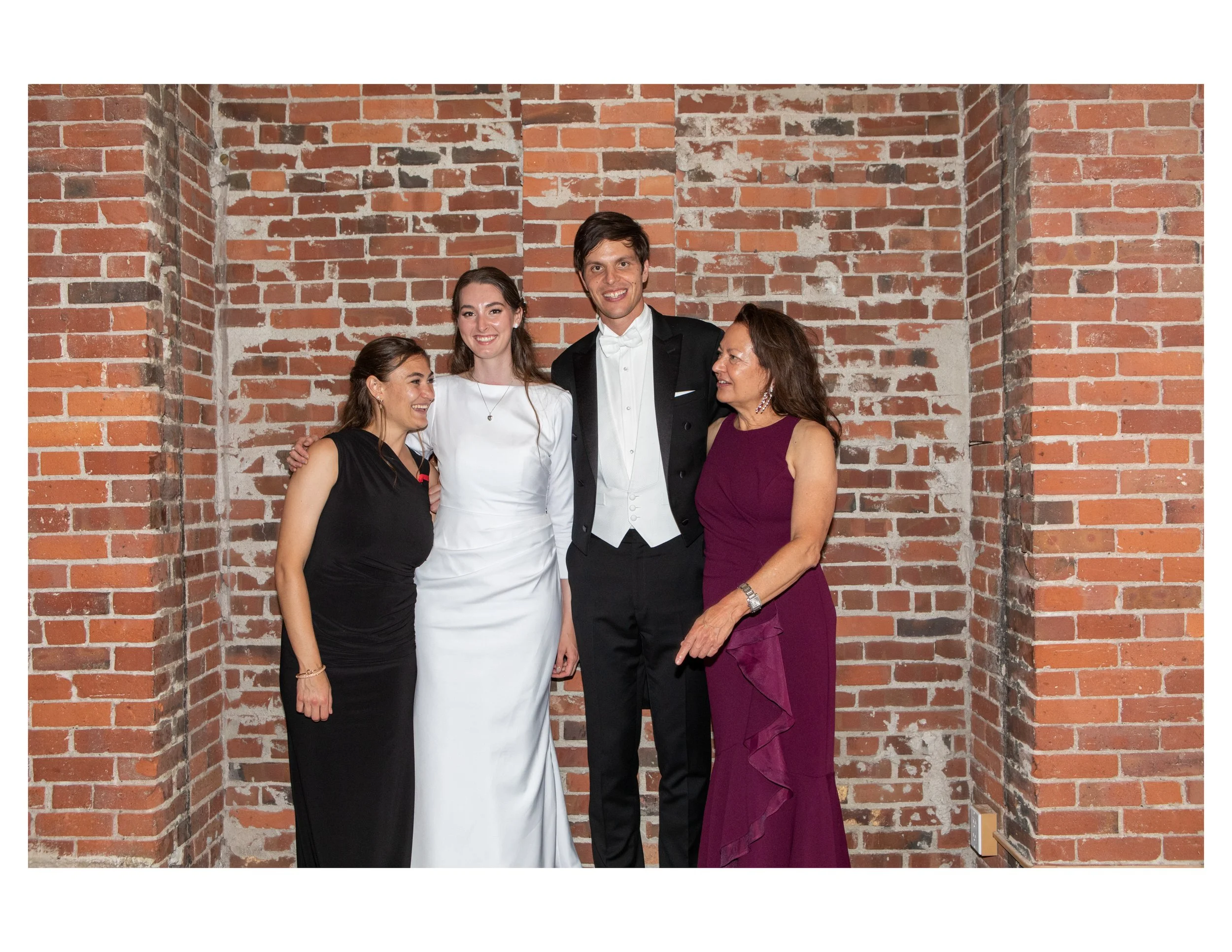 Group of five people dressed in formal attire standing in front of a brick wall, smiling and posing together at a celebration or event.