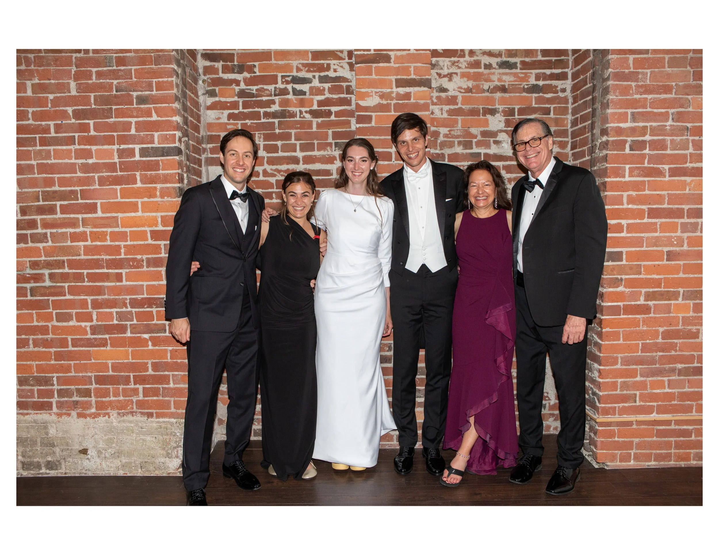 Group of six people dressed in formal attire, standing in front of a brick wall. Four men are in black tuxedos, two women are in elegant dresses, one in black and the other in purple. They are smiling and posing for a photo.
