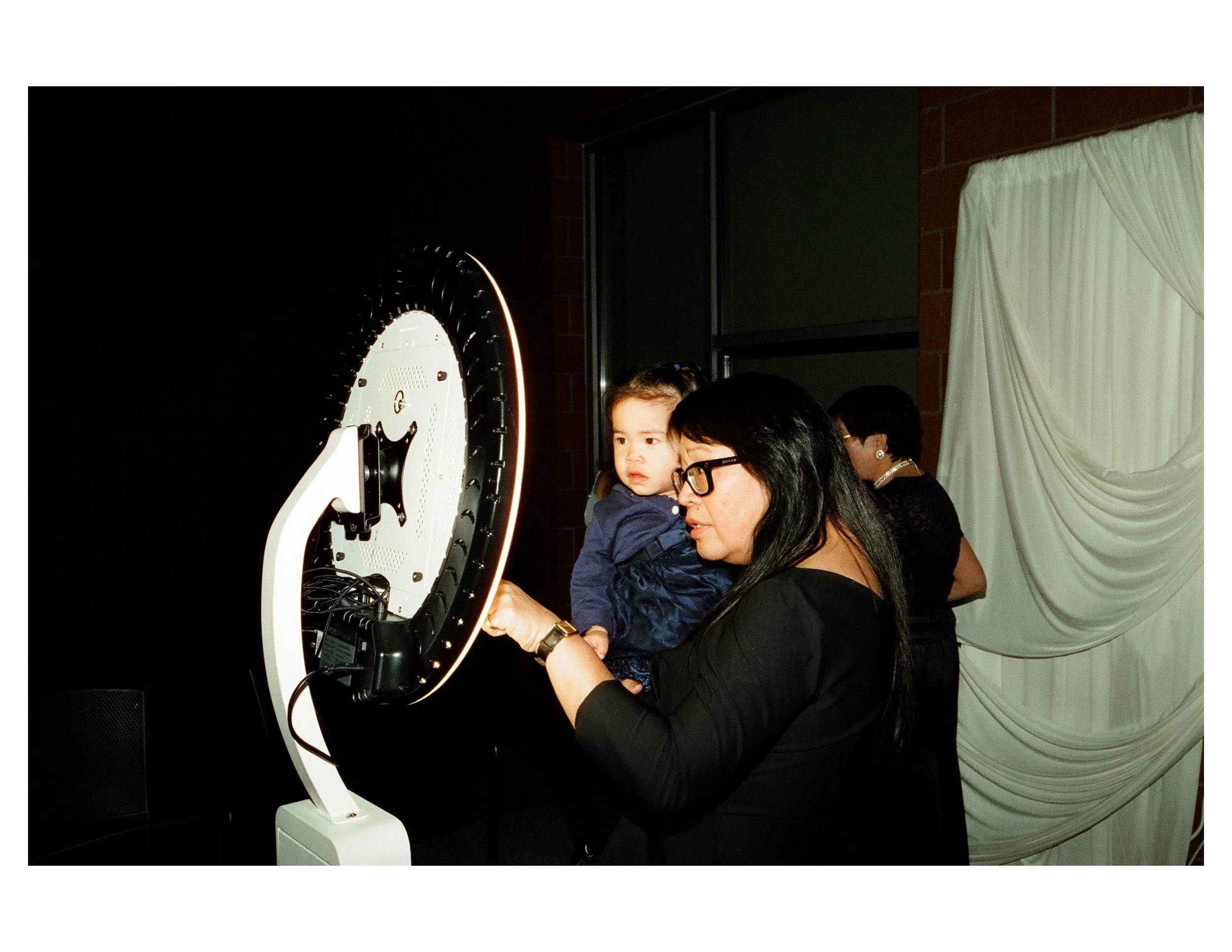 A woman holding a young child while looking at a medical or scientific device with a circular design, set against a dark backdrop with a white curtain in the background.