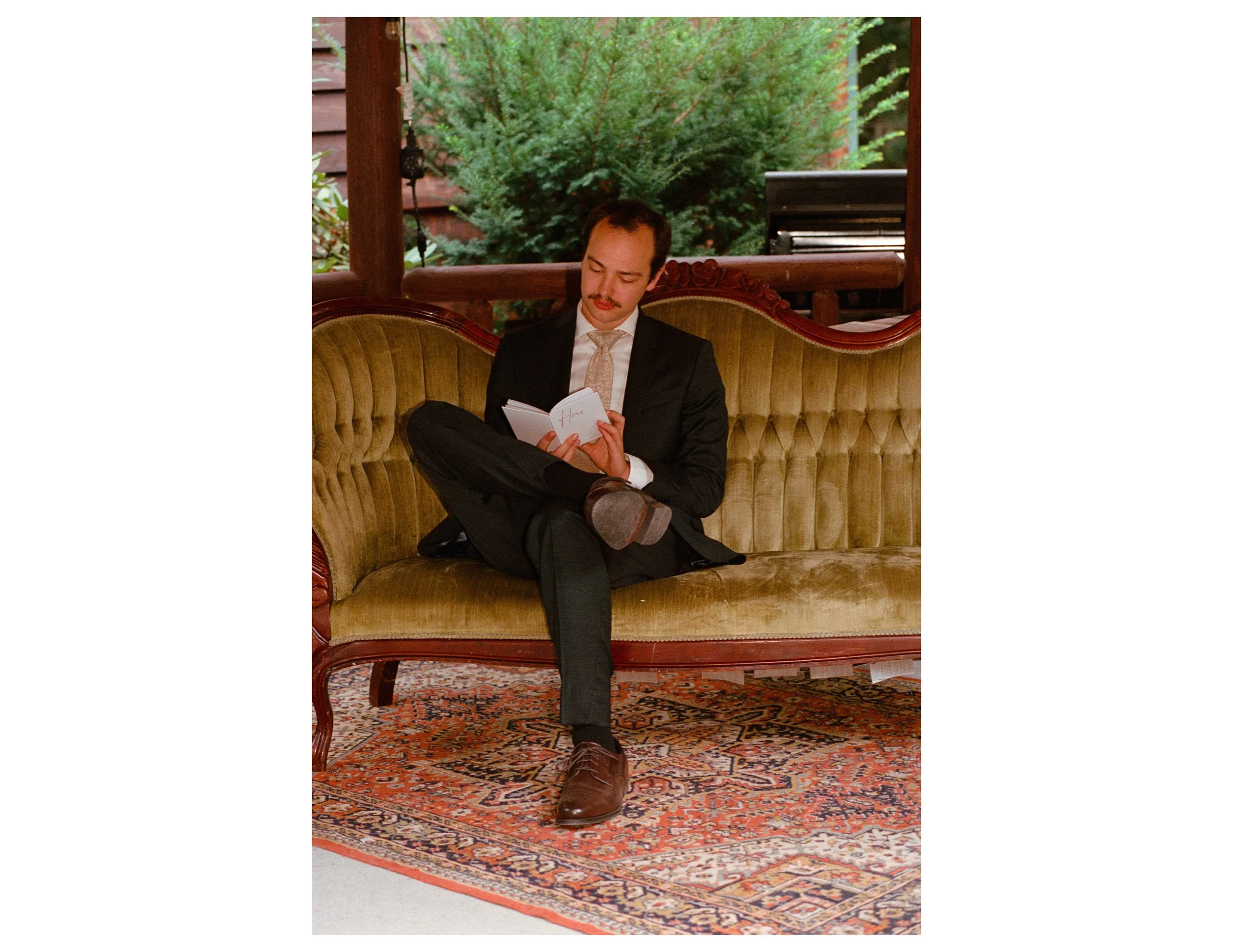 Man with dark hair, mustache, and light skin sitting on a vintage sofa reading a book, dressed in a dark suit and beige tie, in a room with a patterned rug and a large green plant outside the window.