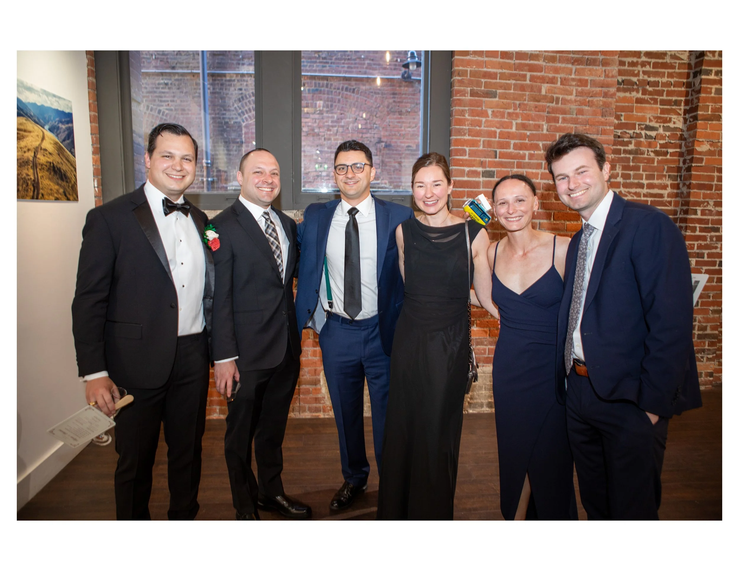 Group of six people dressed in formal attire standing close together inside a brick-walled room with large windows. They are smiling and posing for the photo.