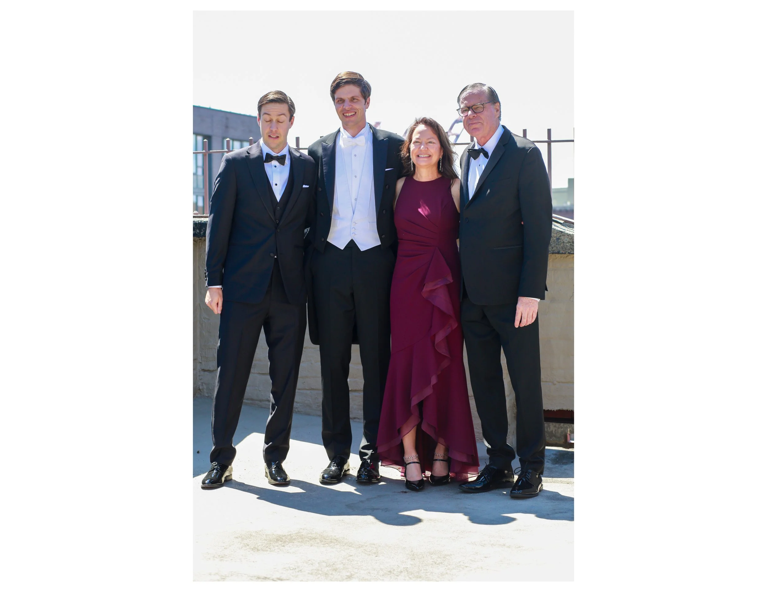 Four people dressed in formal attire posing outdoors on a rooftop or balcony, with buildings and a crane visible in the background.