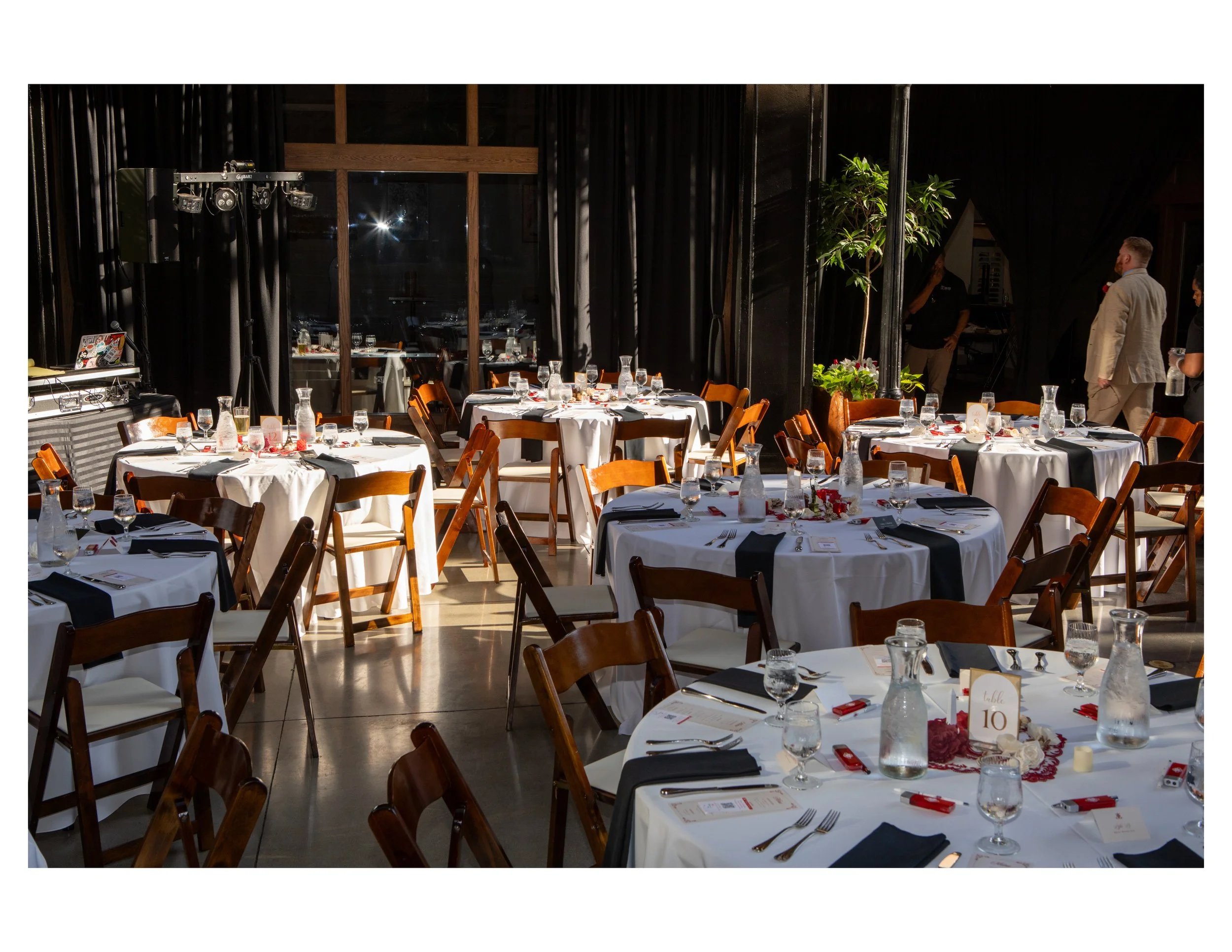 A banquet hall set up for a formal event with round tables, white tablecloths, glassware, and black napkins, with a stage and DJ equipment in the background.
