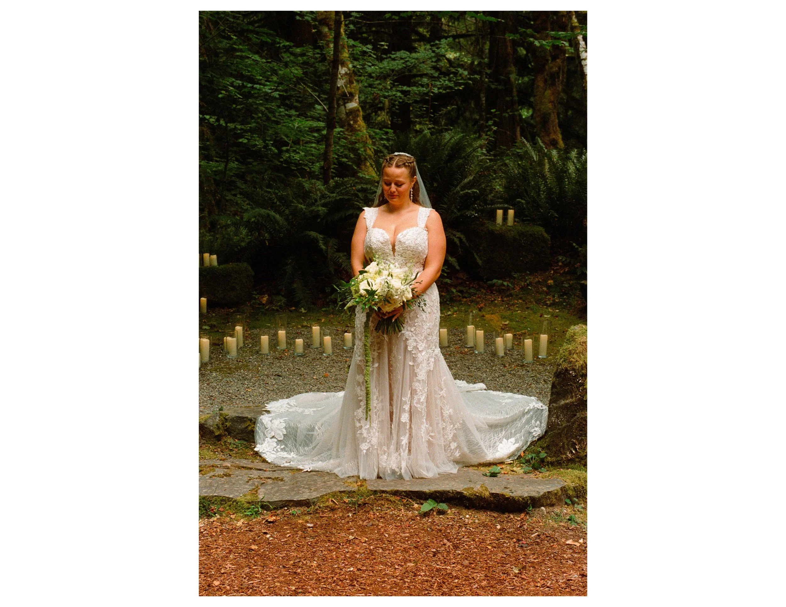 A bride in a white lace wedding dress holding a bouquet of white flowers standing outdoors in a forest with candles on the ground around her.