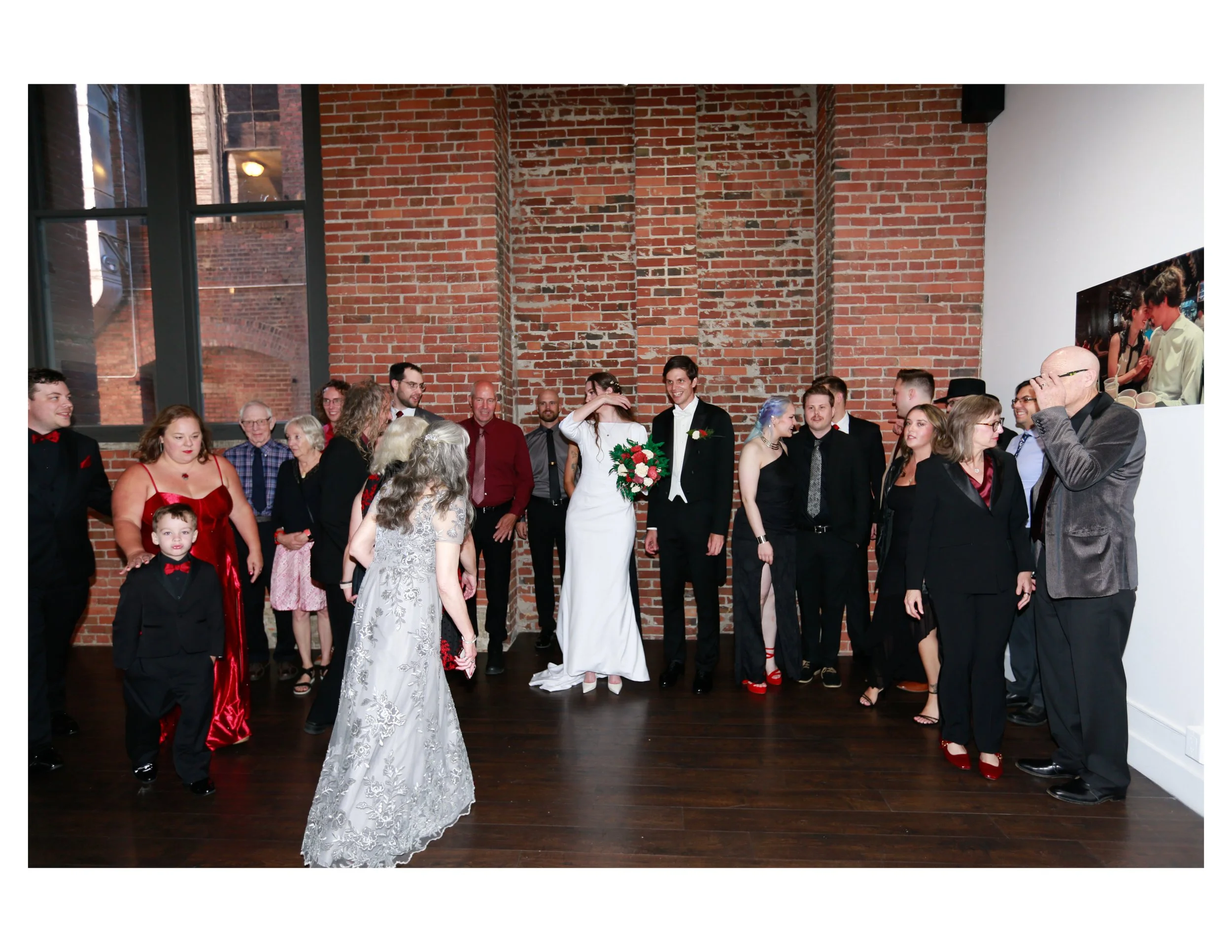 A group of people gathered indoors for a celebration or event, with a brick wall and large windows in the background.