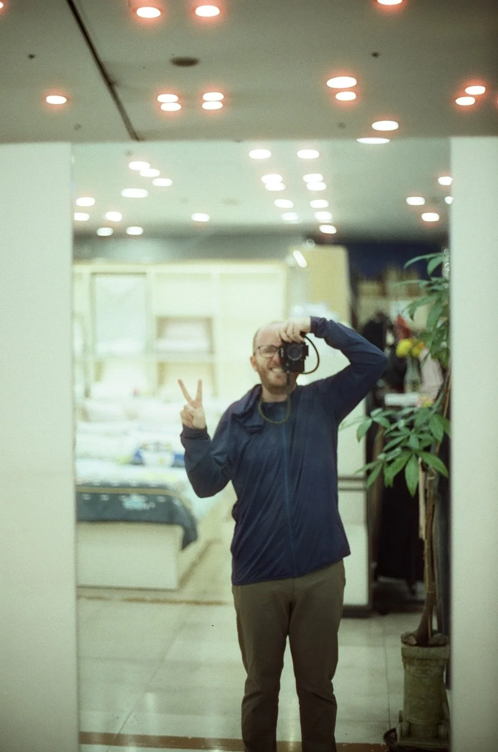 A man taking a selfie in a mirror holding a camera with one hand and making a peace sign with the other. The man is smiling and standing in a room with a bed and a plant.