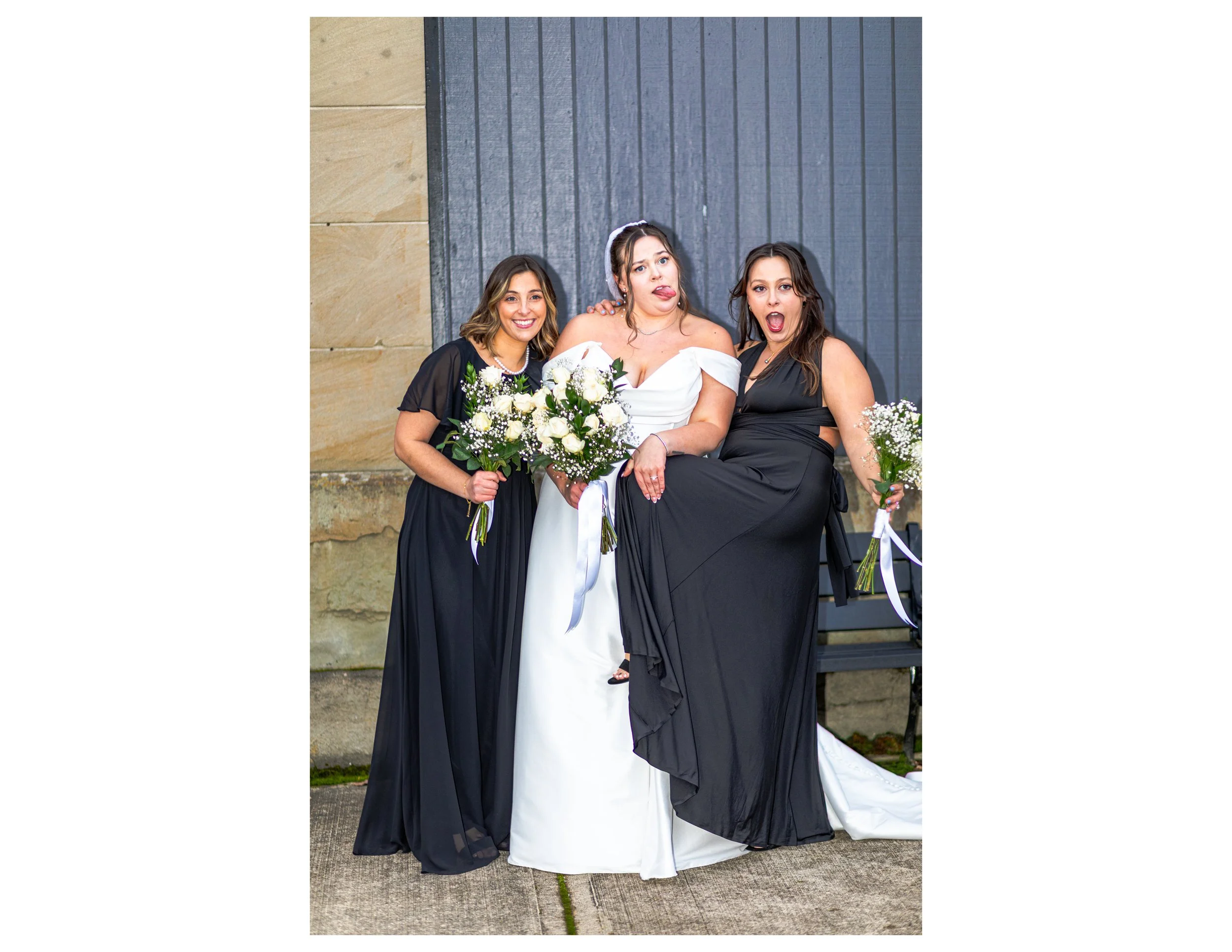 Three women standing together at a wedding, with two women in black dresses holding bouquets of white flowers and the woman in white wearing a wedding dress and veil, making playful expressions.