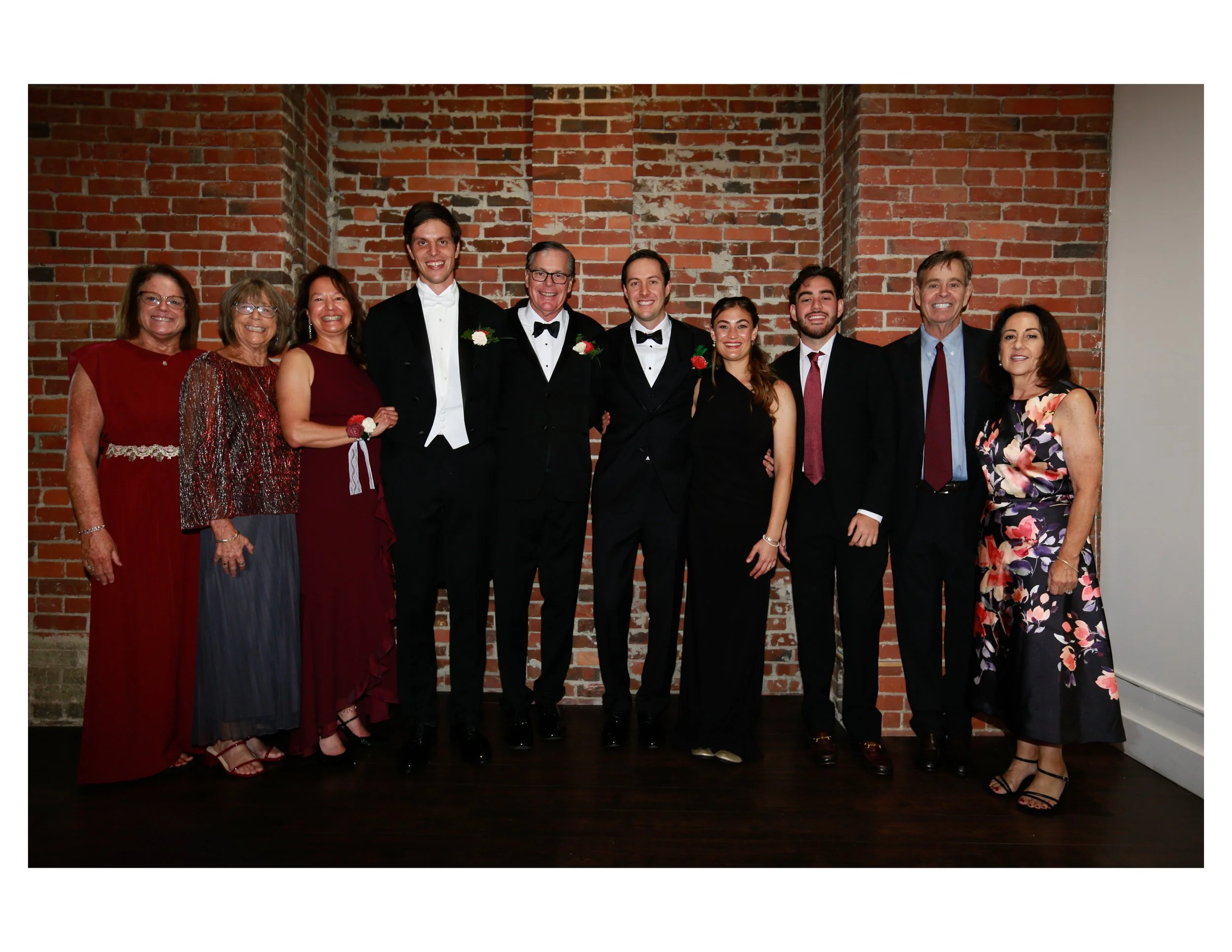 Group of ten people dressed in formal attire standing together in front of a brick wall, smiling at the camera.