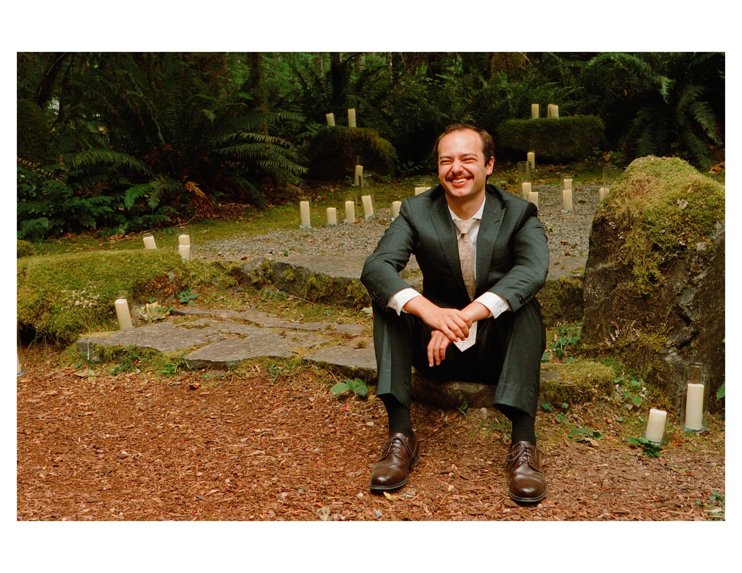 A man in a suit sitting outdoors in a wooded area, smiling and squinting.