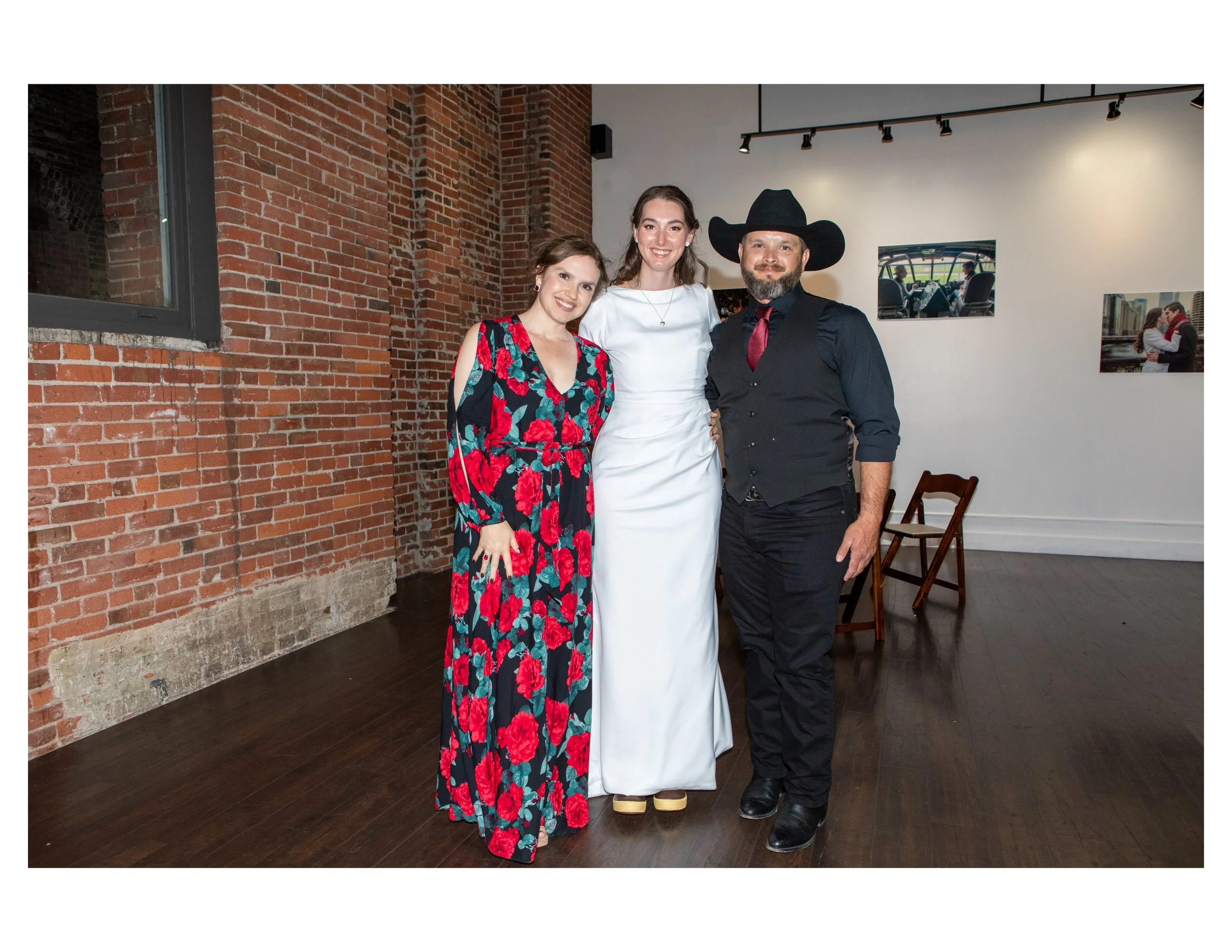 Three people smiling, standing indoors against a brick wall, dressed semi-formally. The woman on the left wears a floral dress, the woman in the center wears a white dress, and the man on the right wears a black cowboy hat, black vest, red tie, and b