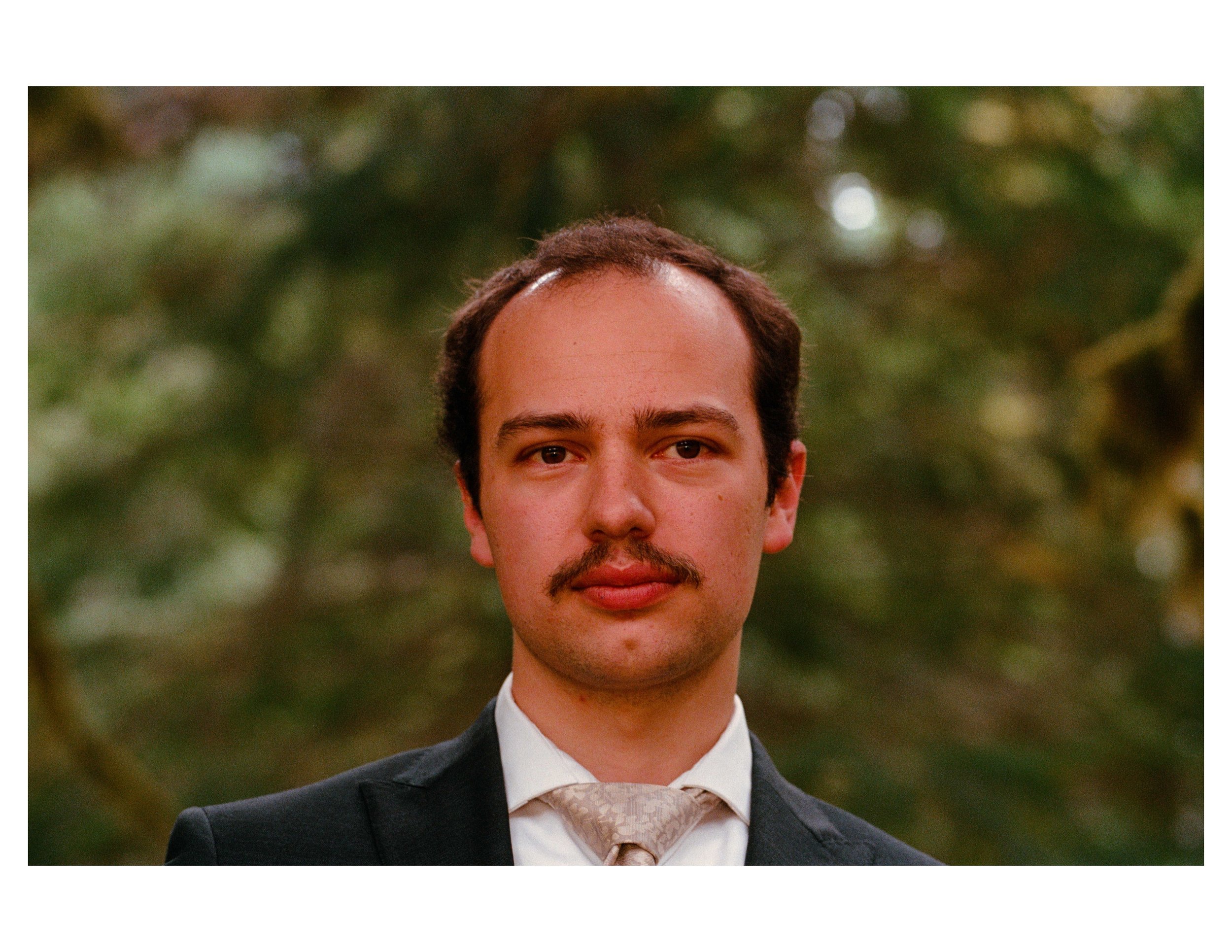 Close-up of a young man in a suit and tie, outdoors with blurred greenery in the background.
