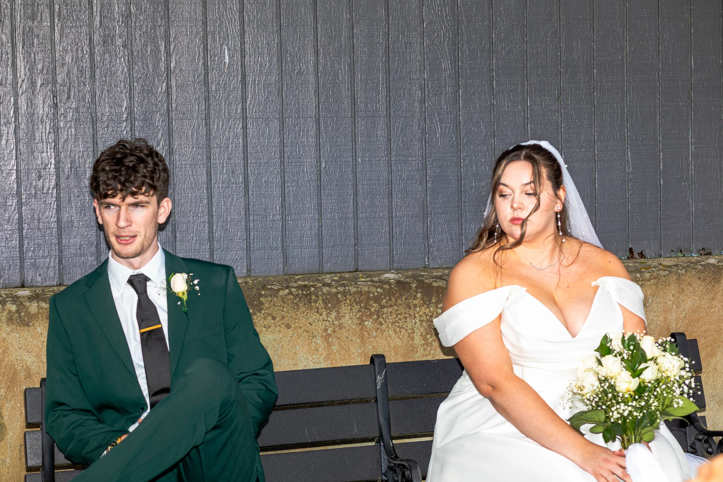 A young man in a green suit with a white shirt and black tie sitting on a bench, and a young woman in a white wedding dress holding a bouquet of white flowers sitting next to him, both in front of a dark wooden panel.