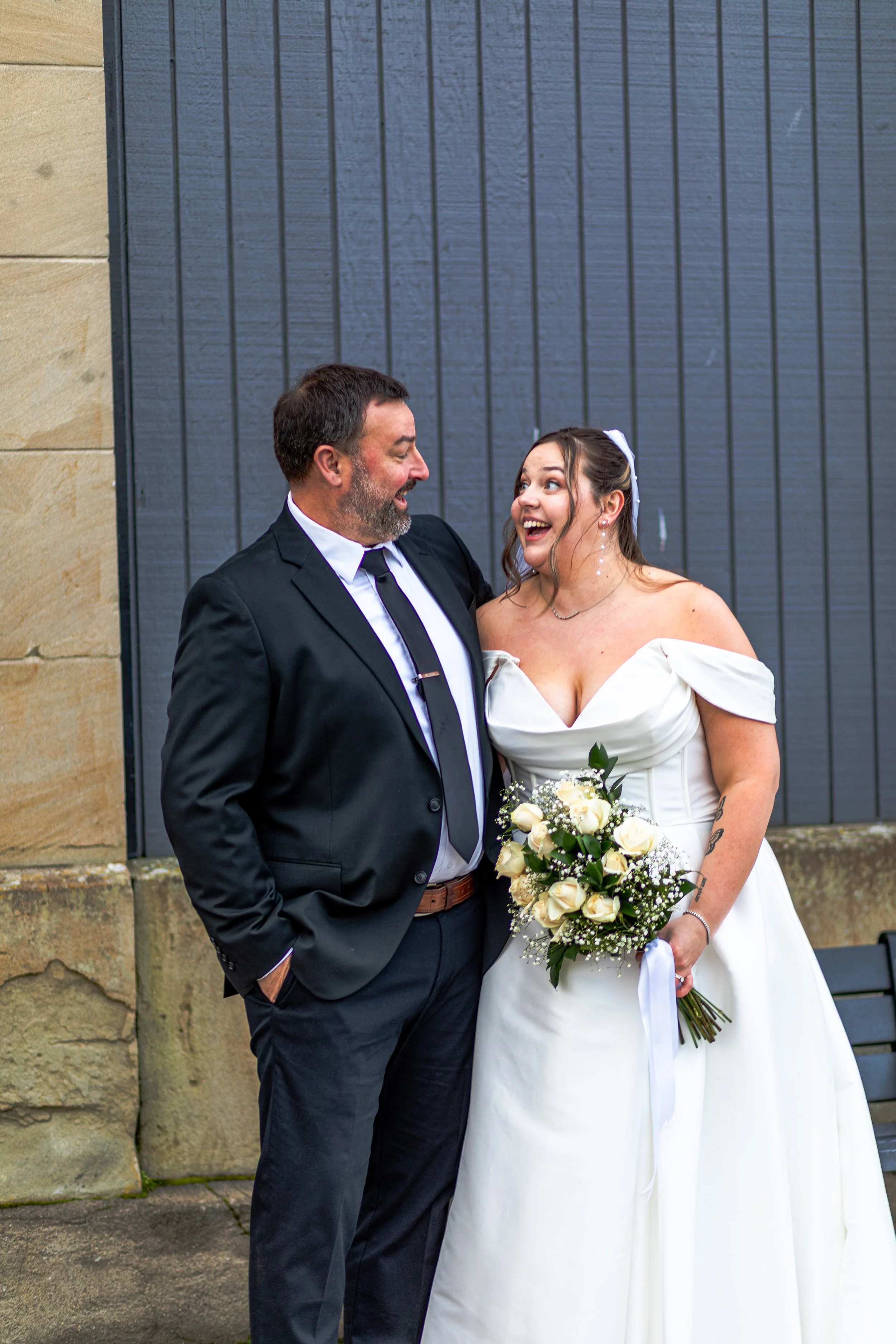 A bride and groom sharing a joyful moment outdoors, with the bride holding a bouquet of white roses and baby's breath, standing in front of a blue and beige wall.