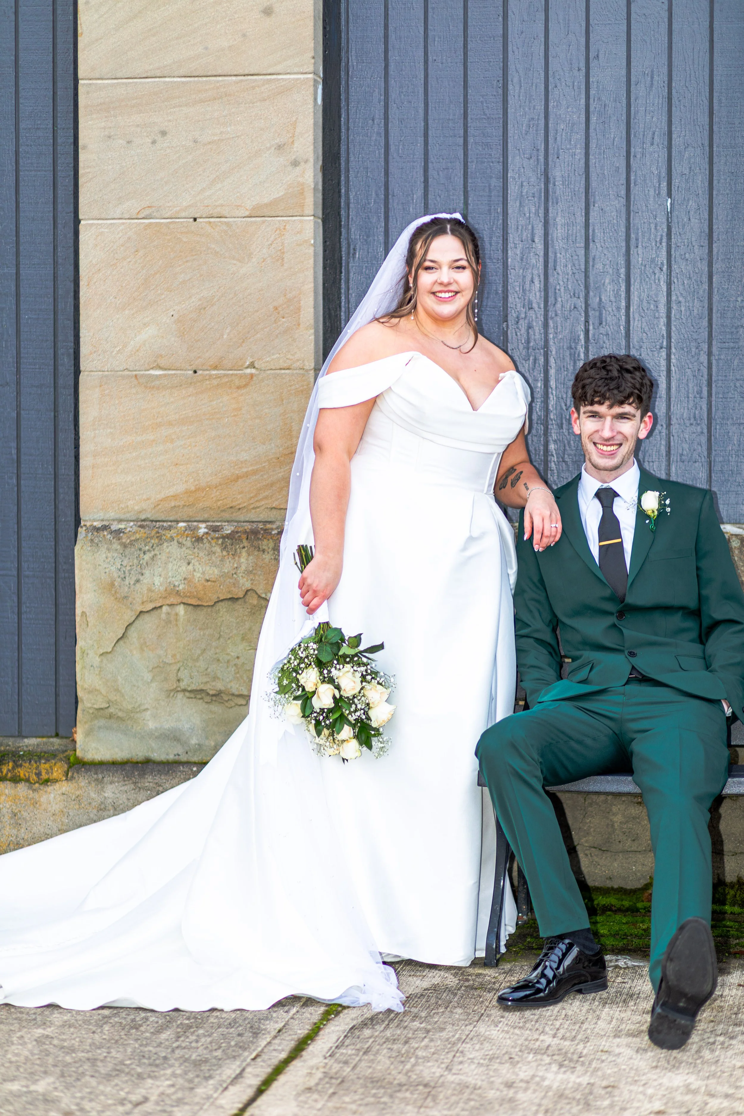 A bride in a white off-shoulder wedding dress holding a bouquet of white roses, standing next to a seated groom in a dark green suit with a white shirt, black tie, and boutonniere, against a dark wood and stone wall background.