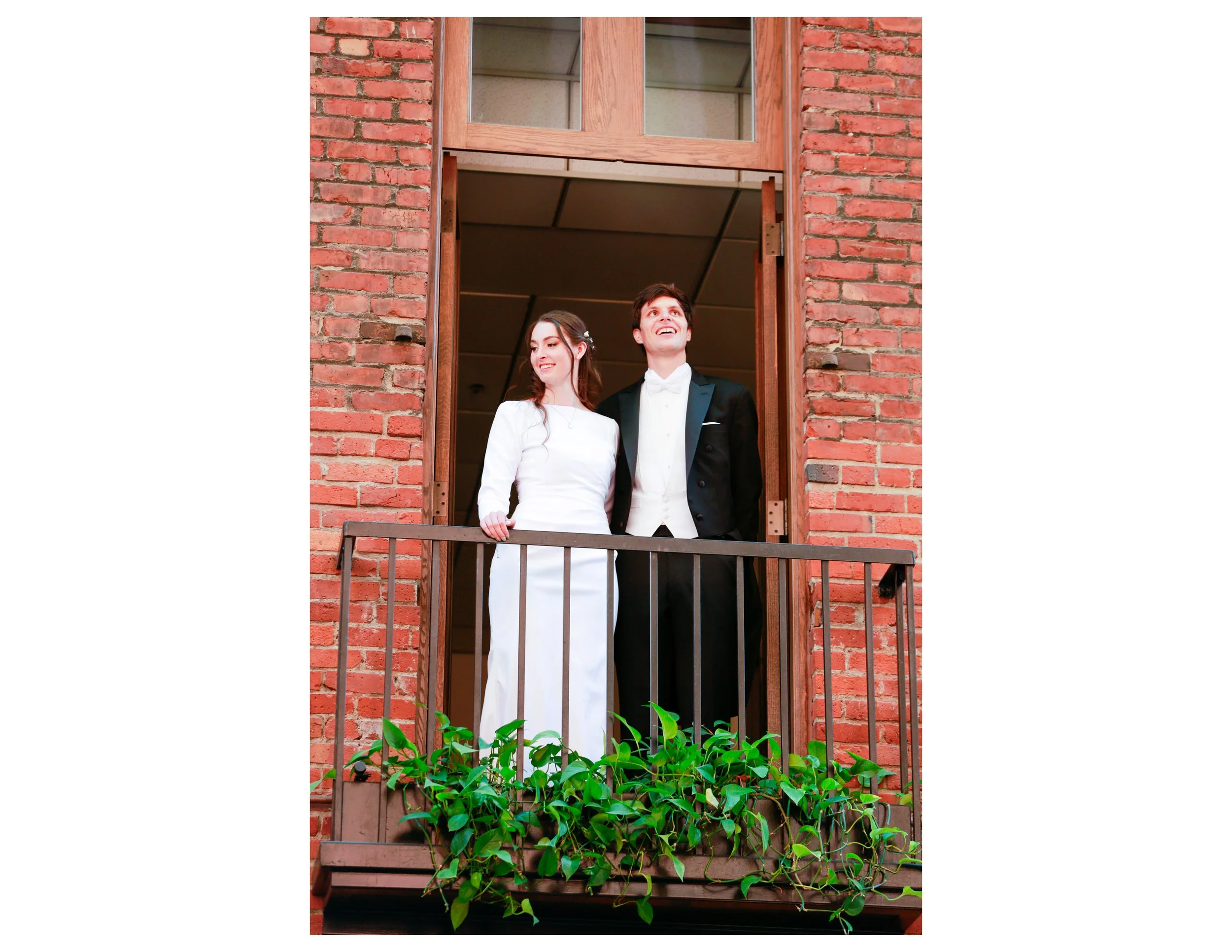 Two people, a woman in a white dress and a man in a tuxedo, are standing on a balcony looking up and smiling, with a brick wall and window behind them and green plants in the foreground.
