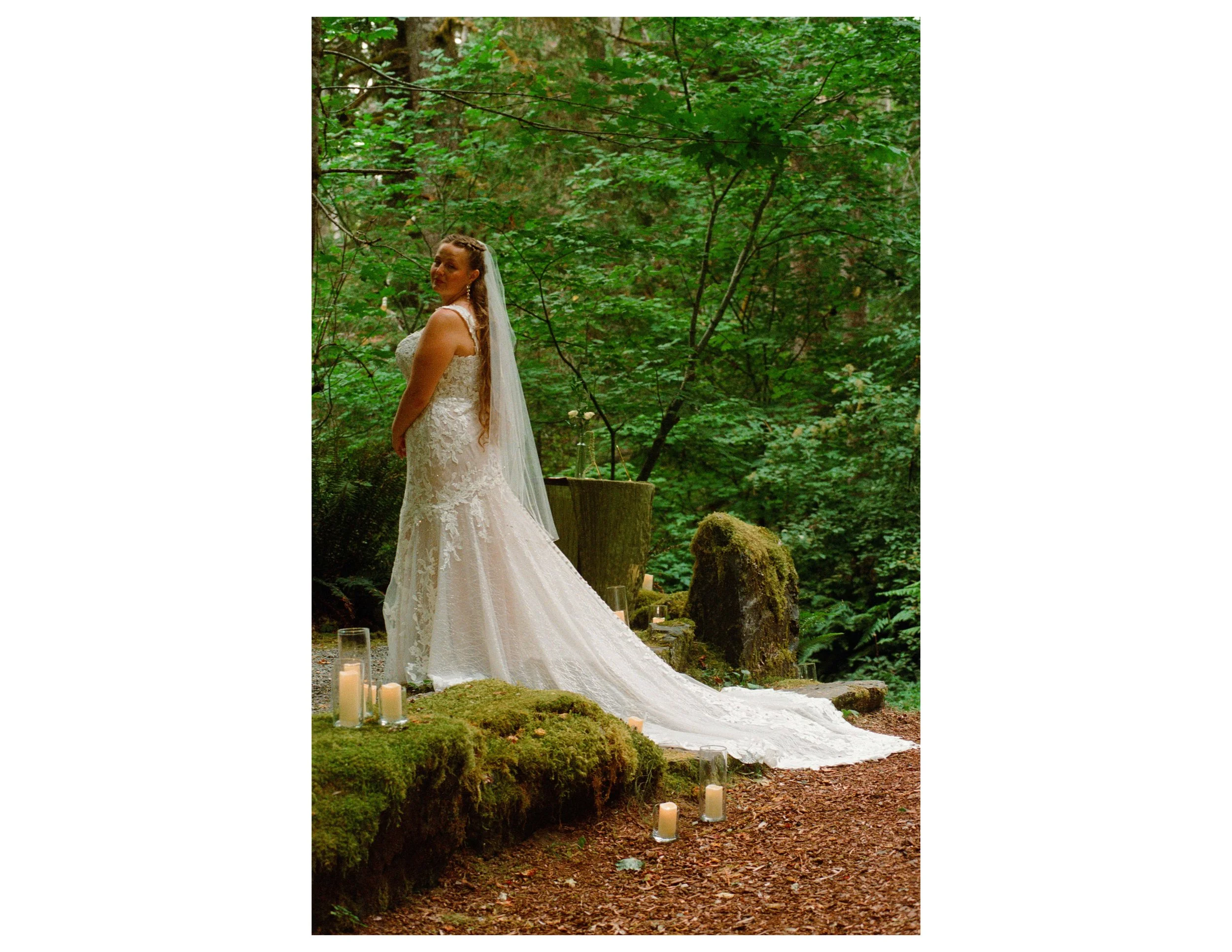 A bride in a lace wedding dress and veil stands in a forested outdoor setting, surrounded by trees and candles placed on the ground.