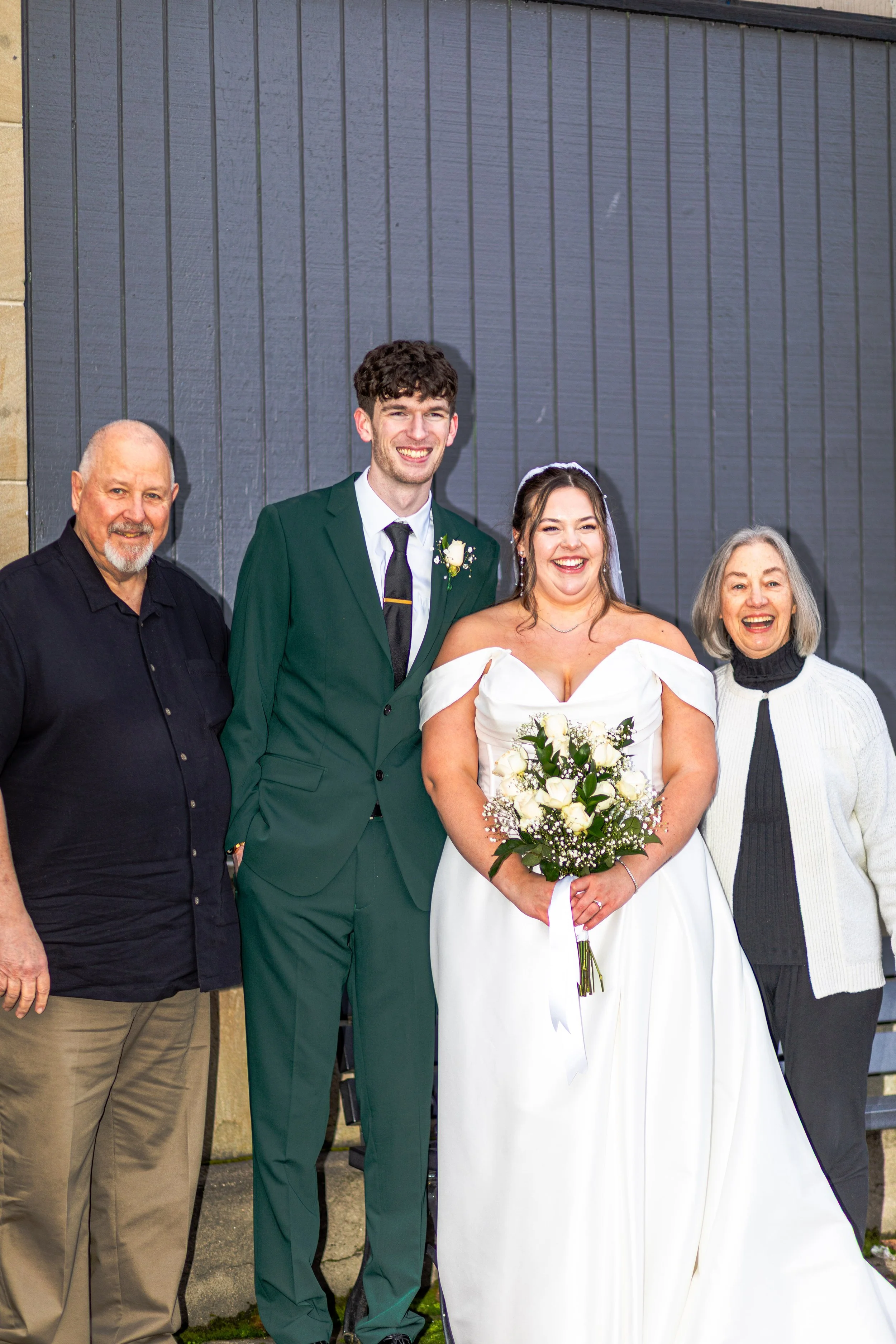 Group of people at a wedding, including bride holding a bouquet of white roses, groom in a green suit, and two older adults standing beside them, smiling outside in front of a dark wooden background.