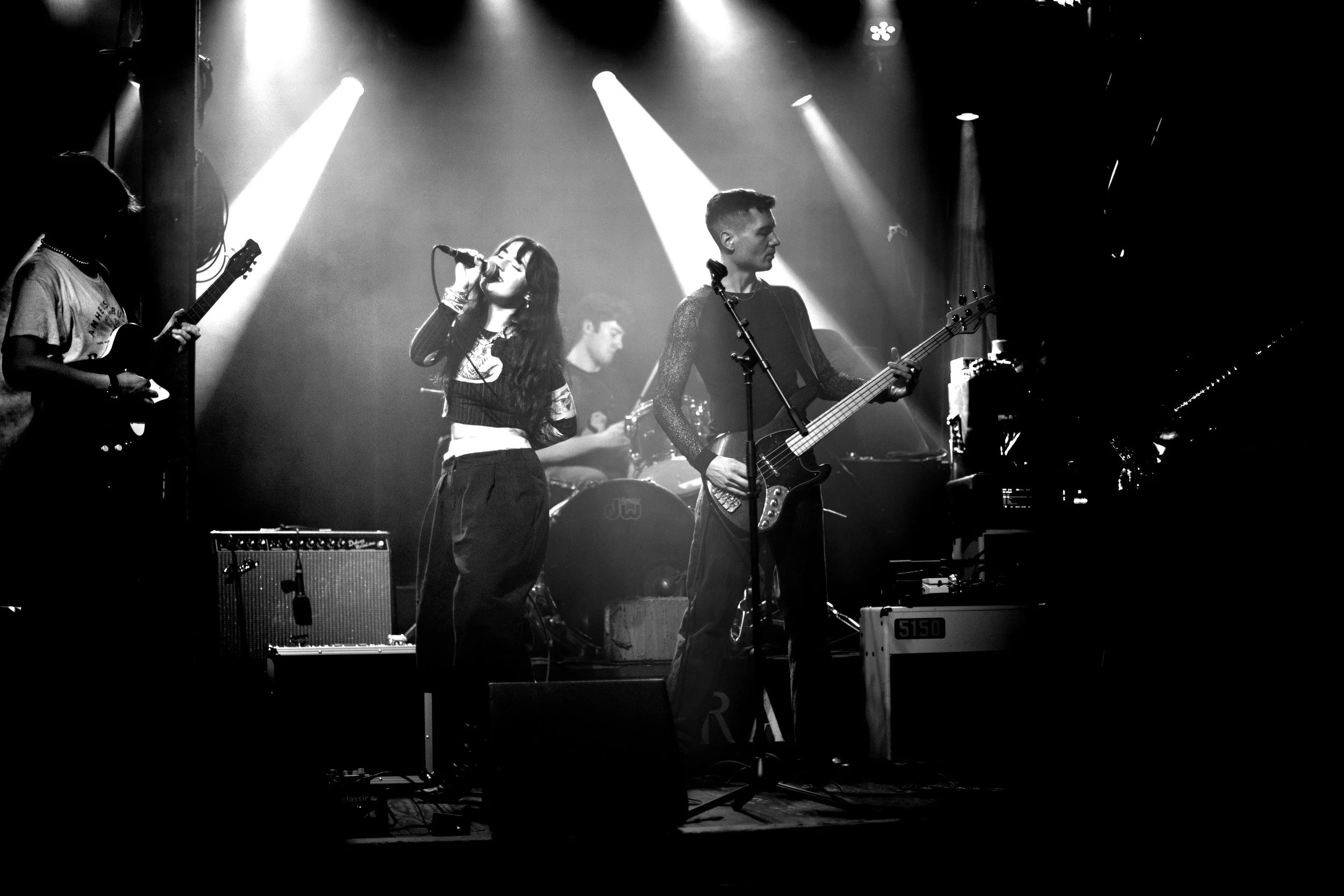 Black and white photo of a band performing on stage, with a female singer in the center singing into a microphone, a male guitarist on her right playing an electric guitar, and a drummer in the background. Stage lights illuminate the scene.