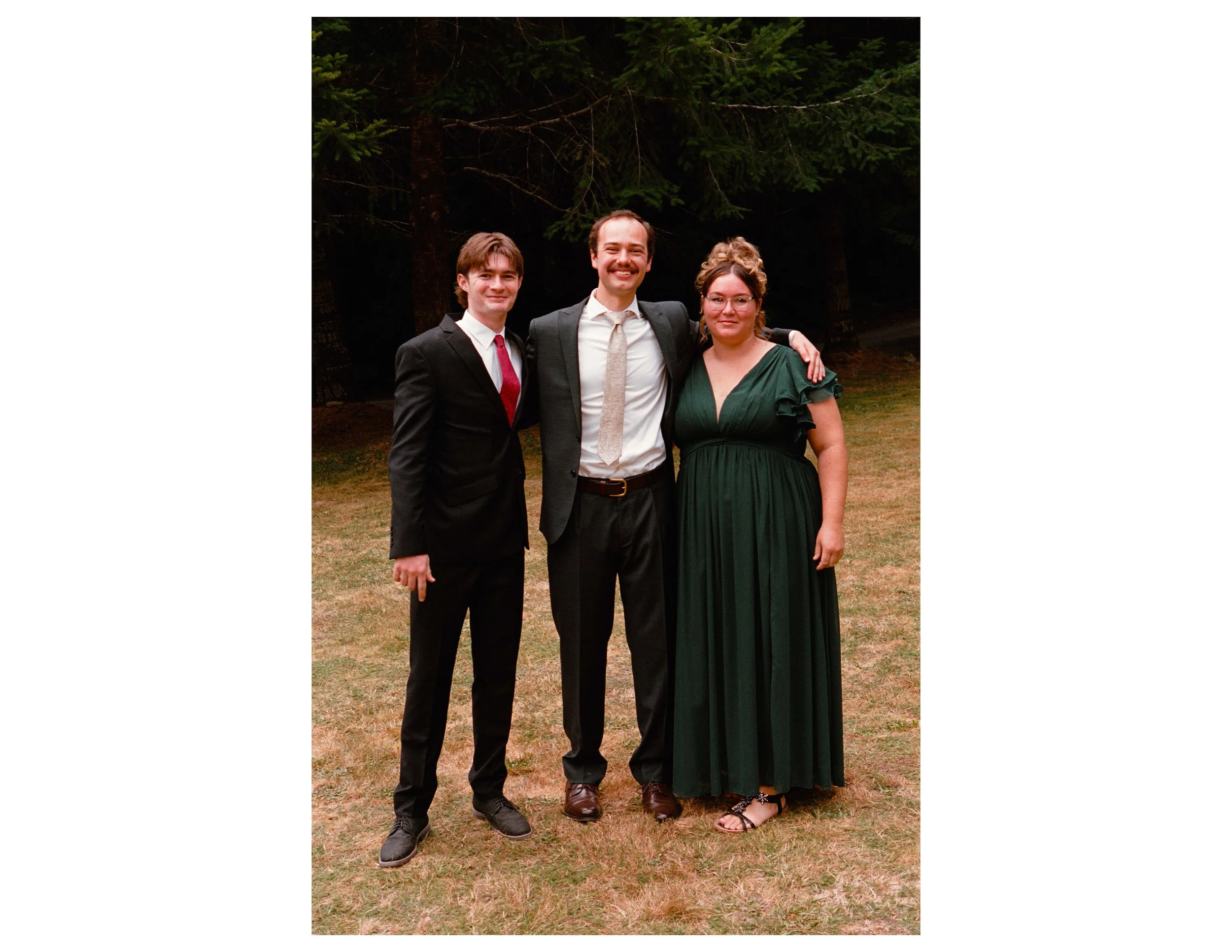 Three people dressed in formal attire standing outdoors with trees in the background, smiling at the camera.