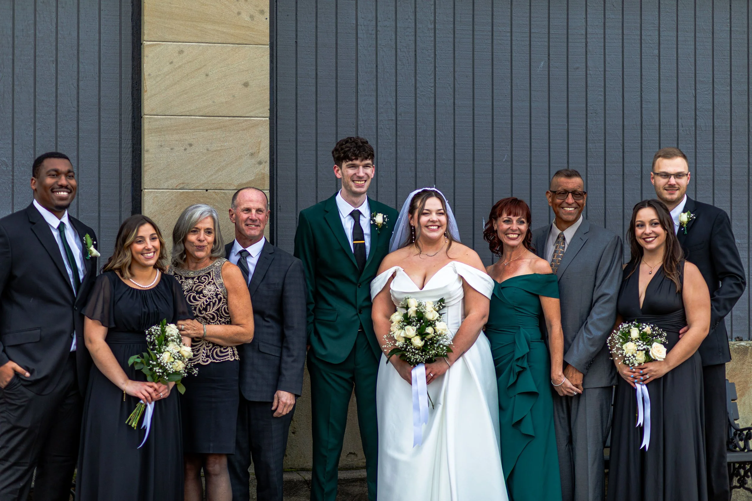 Group of ten people dressed in wedding attire, standing outdoors in front of a building, celebrating a wedding.