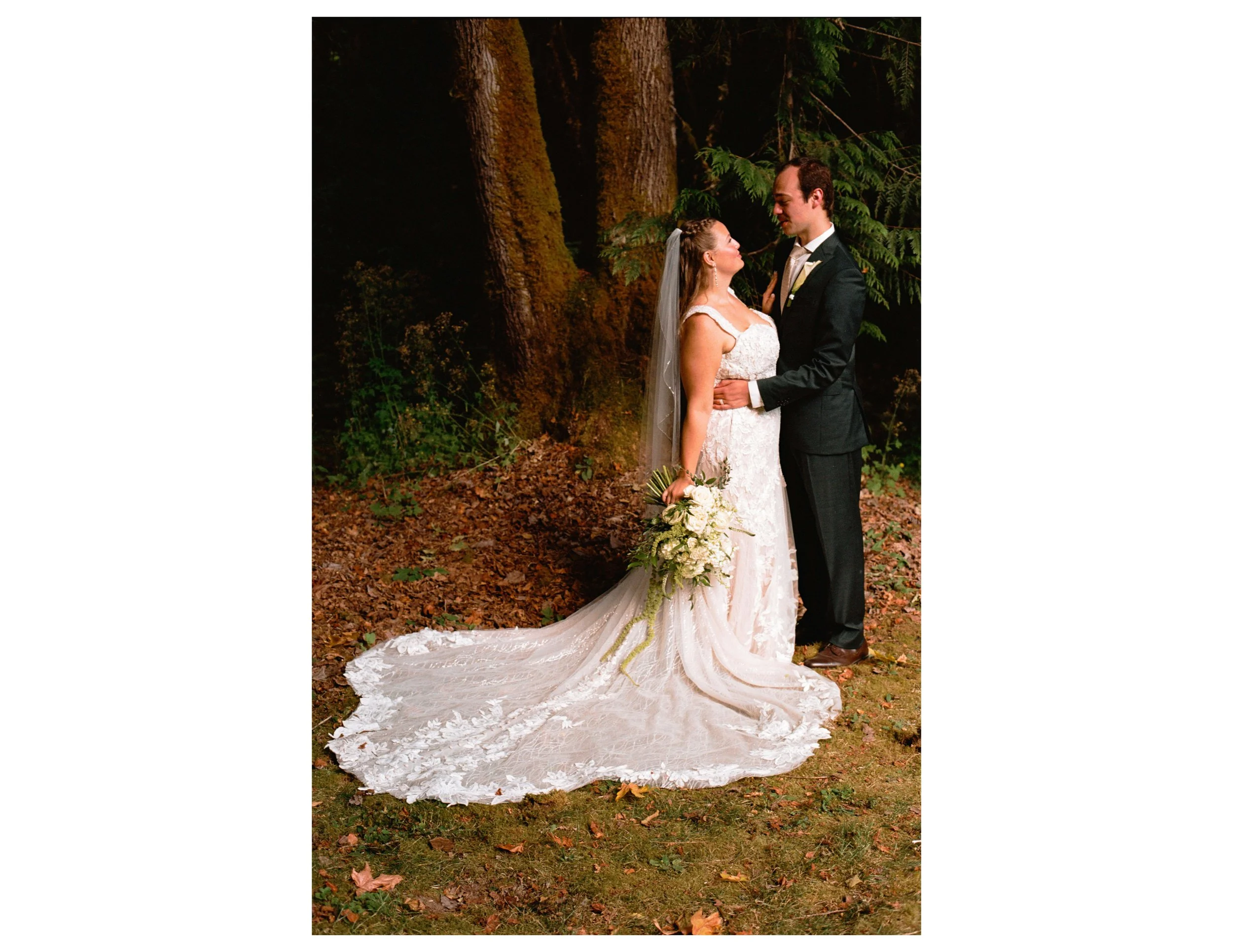 A bride and groom standing in a wooded area during a wedding photoshoot. The bride wears a white lace wedding gown with a long train and holds a bouquet of white flowers. The groom is dressed in a dark tuxedo. They are gazing into each other's eyes, 