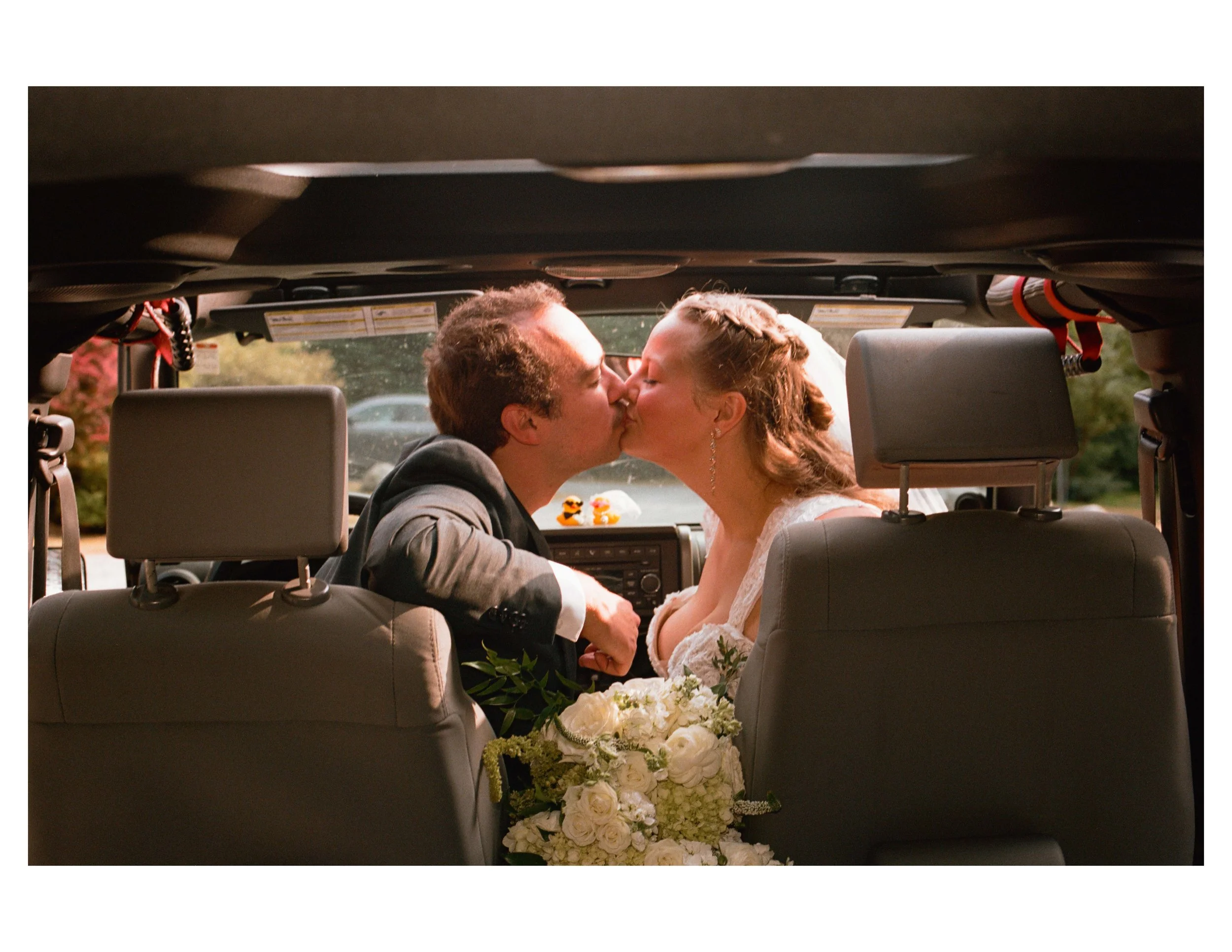 A couple sharing a kiss in the backseat of a car, with a wedding bouquet of white roses and greenery visible.