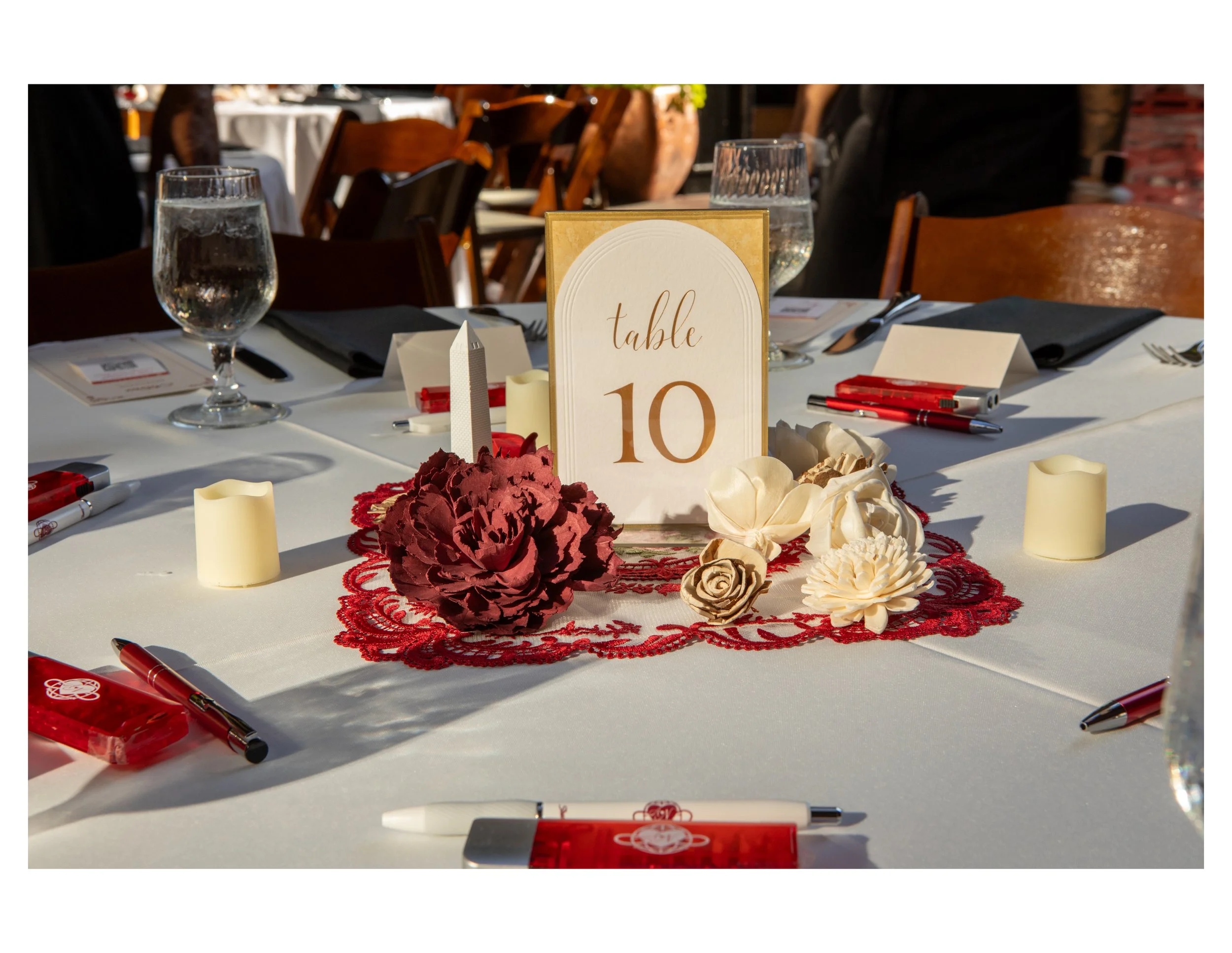 A table set for a celebration with a sign indicating it is table 10, surrounded by flowers, candles, water glasses, and red and white pens.