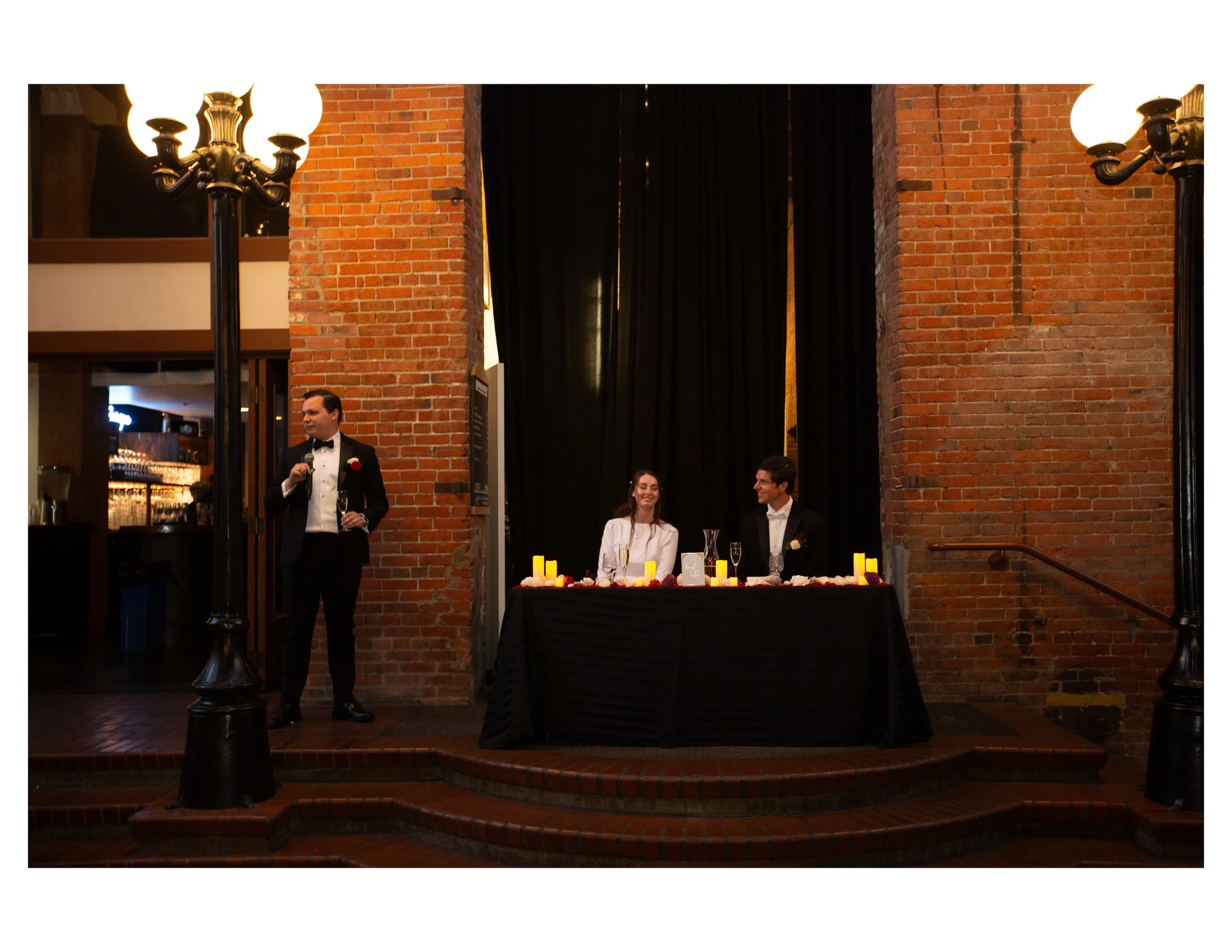 A wedding reception with a couple sitting at a table with candles and floral decorations, a man in a tuxedo holding a glass, and a man in a tuxedo standing nearby; the background features brick walls, black curtains, and vintage street lamps with lit