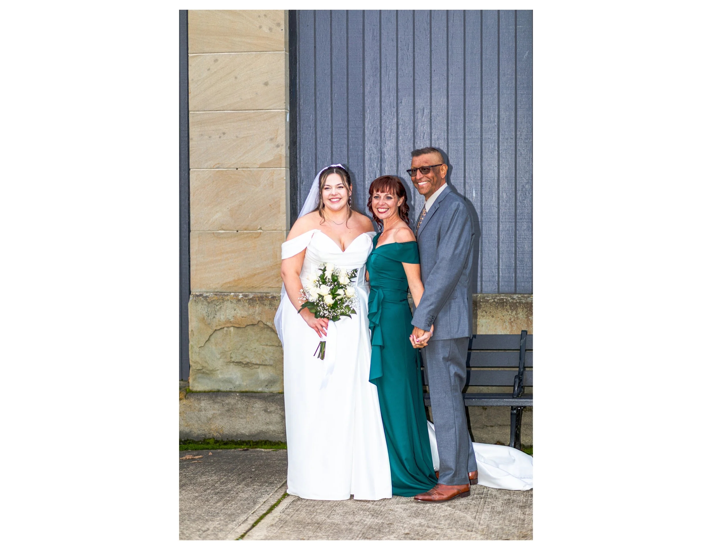 A bride in a white wedding dress holding a bouquet stands next to a woman in a teal dress and a man in a gray suit and glasses, all smiling outside against a blue wooden wall.