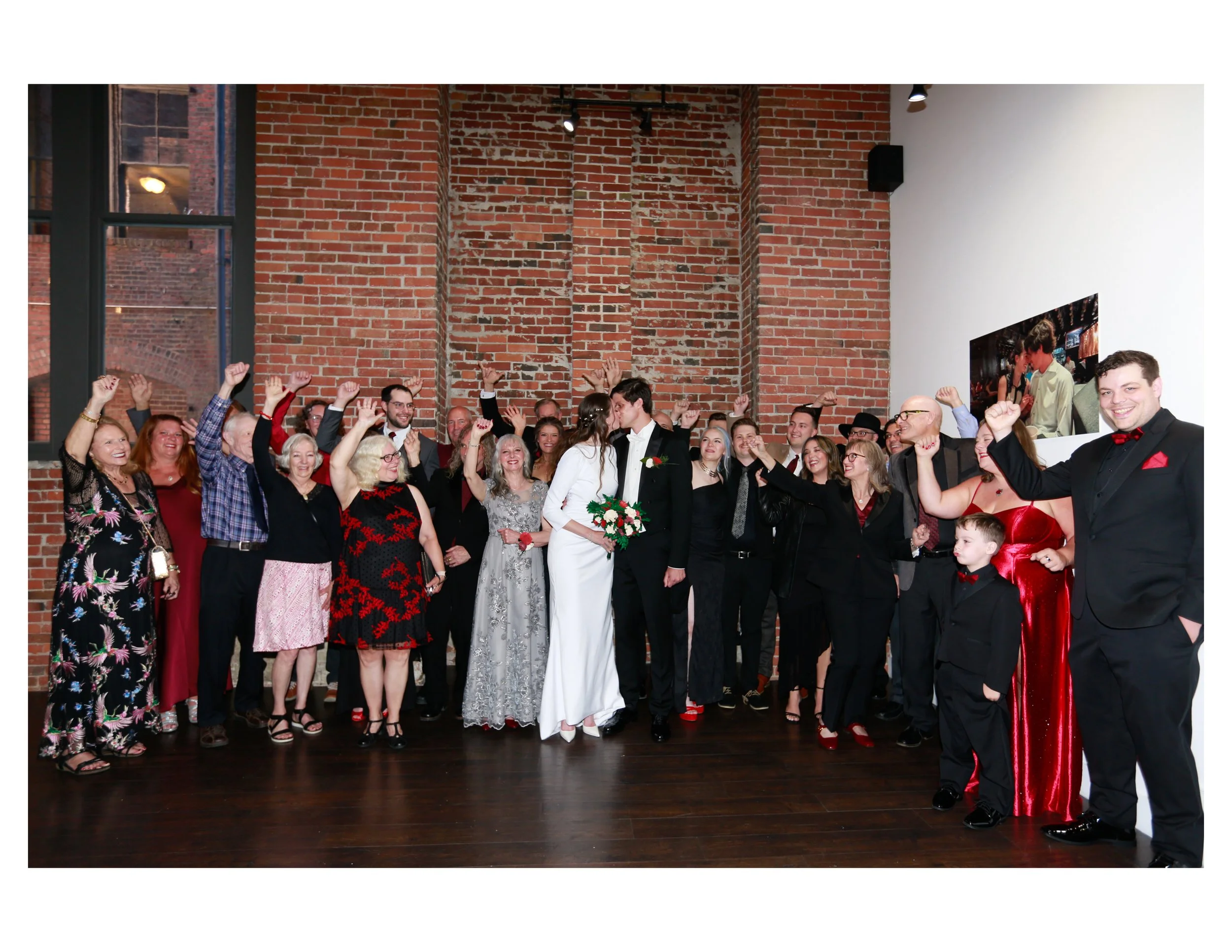 Group of people at a celebration or wedding, smiling and raising their hands, with a bride and groom in the center, in a room with brick walls and a wooden floor.