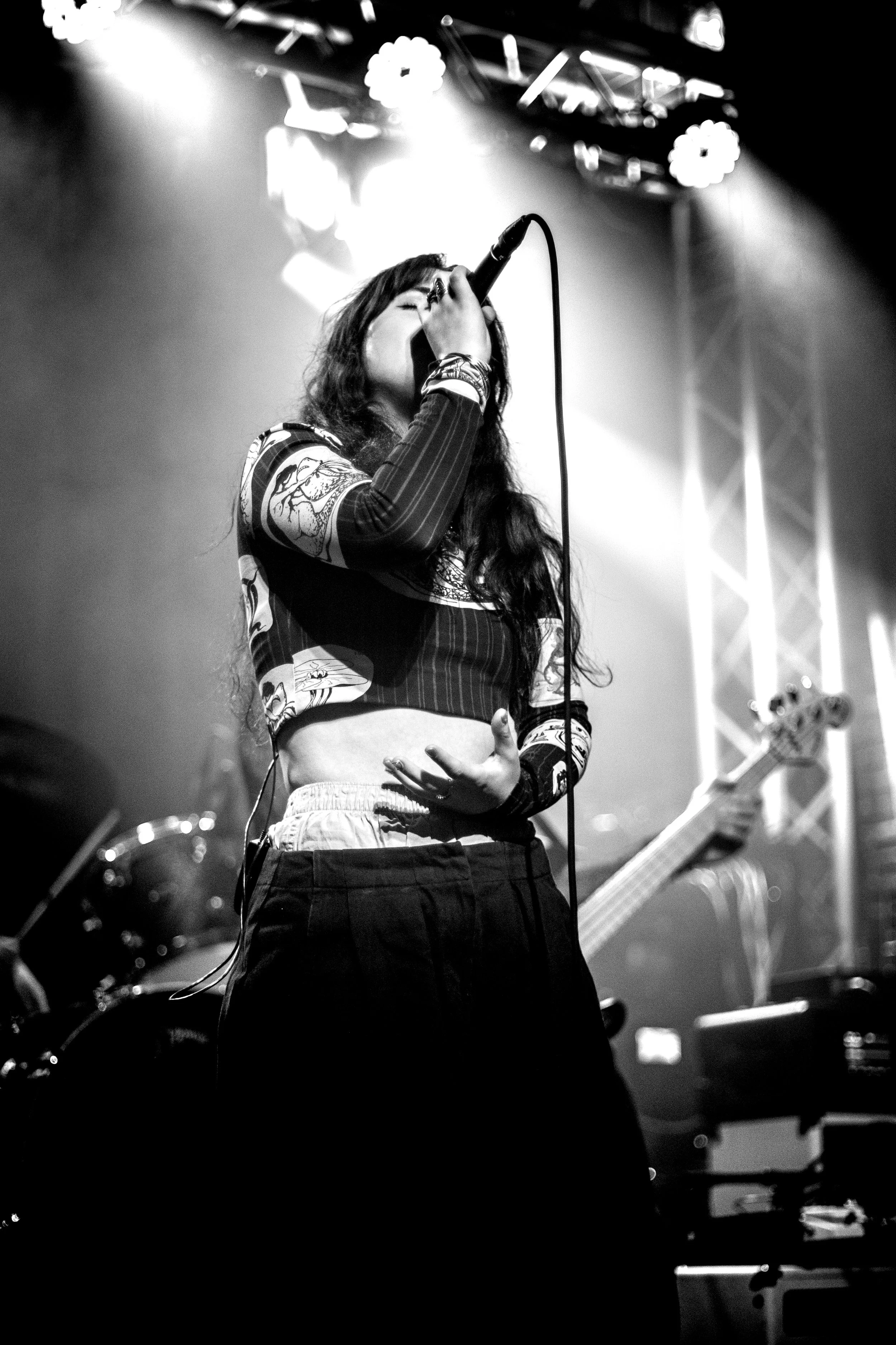 A female singer with long dark hair wearing a patterned long-sleeve crop top and loose pants, singing into a microphone on stage with bright lights and musical instruments in the background.
