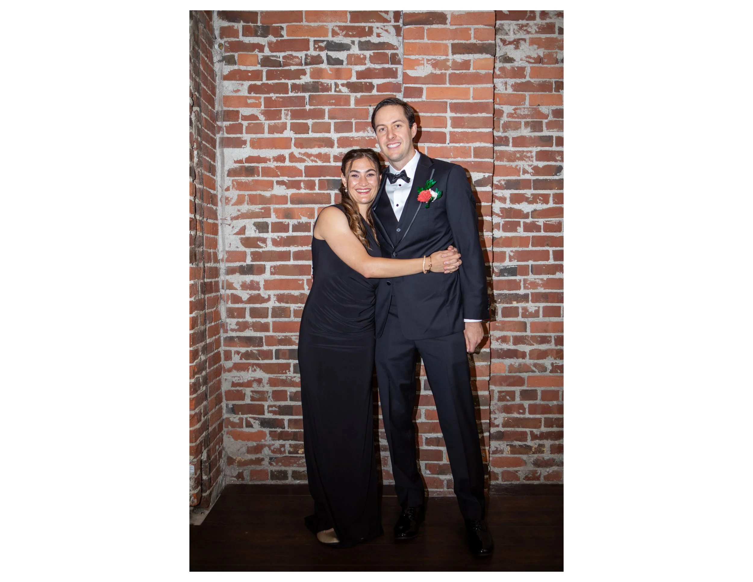 A couple dressed in formal attire standing in front of an exposed brick wall, smiling and embracing.