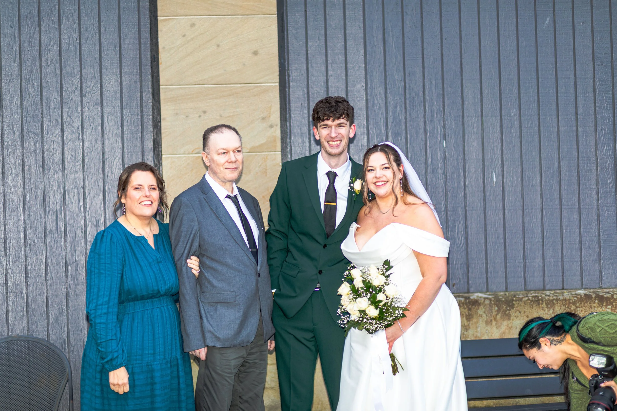 A group of five people at a wedding, including the bride in a white off-the-shoulder gown holding a bouquet, the groom in a green suit with a white boutonniere, and three other individuals dressed in formal attire, posing and smiling in front of a gr